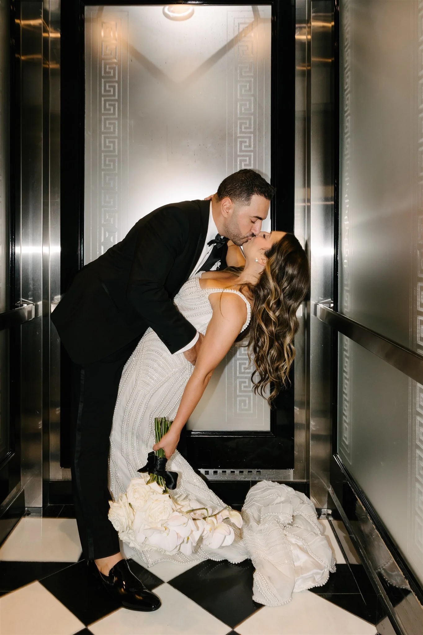 A groom dips and kisses a bride inside an elevator, with the bride holding a bouquet of white flowers, both dressed in wedding attire.