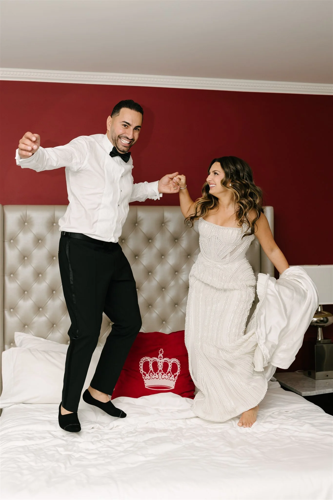 A couple dancing on a bed, with the woman in a white dress and the man in a tuxedo. They are smiling and holding hands, in a room with a red wall and a tufted headboard.