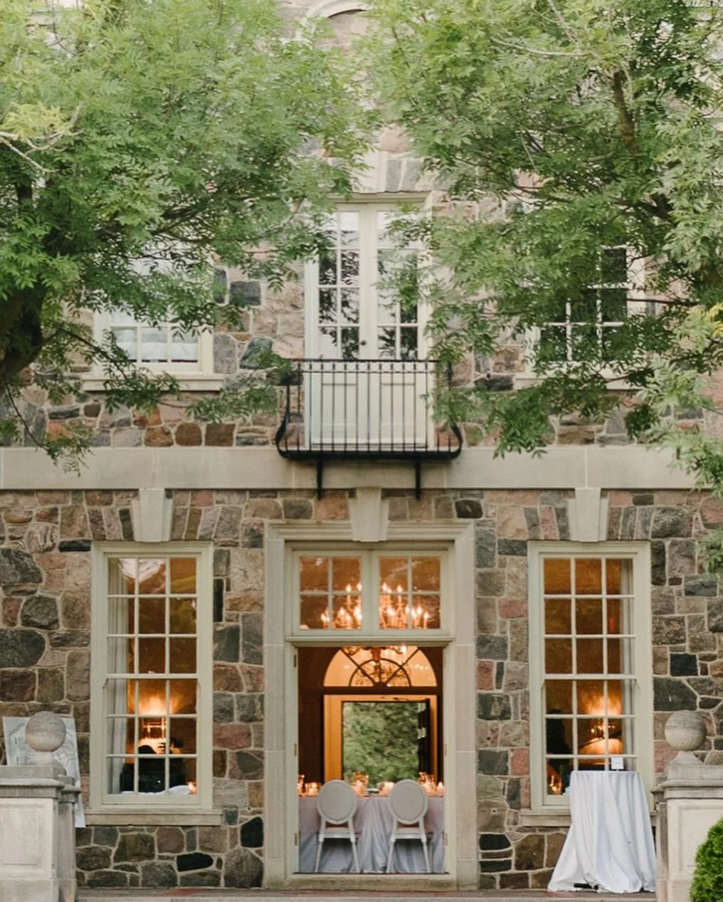 A stone house with large windows and a small balcony on the second floor surrounded by green trees. The ground floor has a doorway with a table set for a meal visible inside, illuminated by warm lighting and a chandelier.