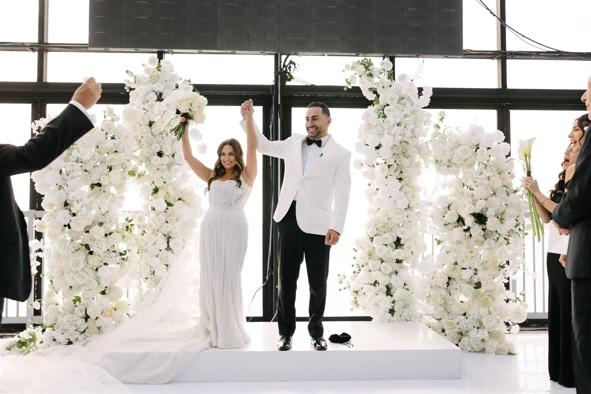 A newly married couple on their wedding day, holding hands and smiling at a ceremony with white floral decorations and guests in formal attire.