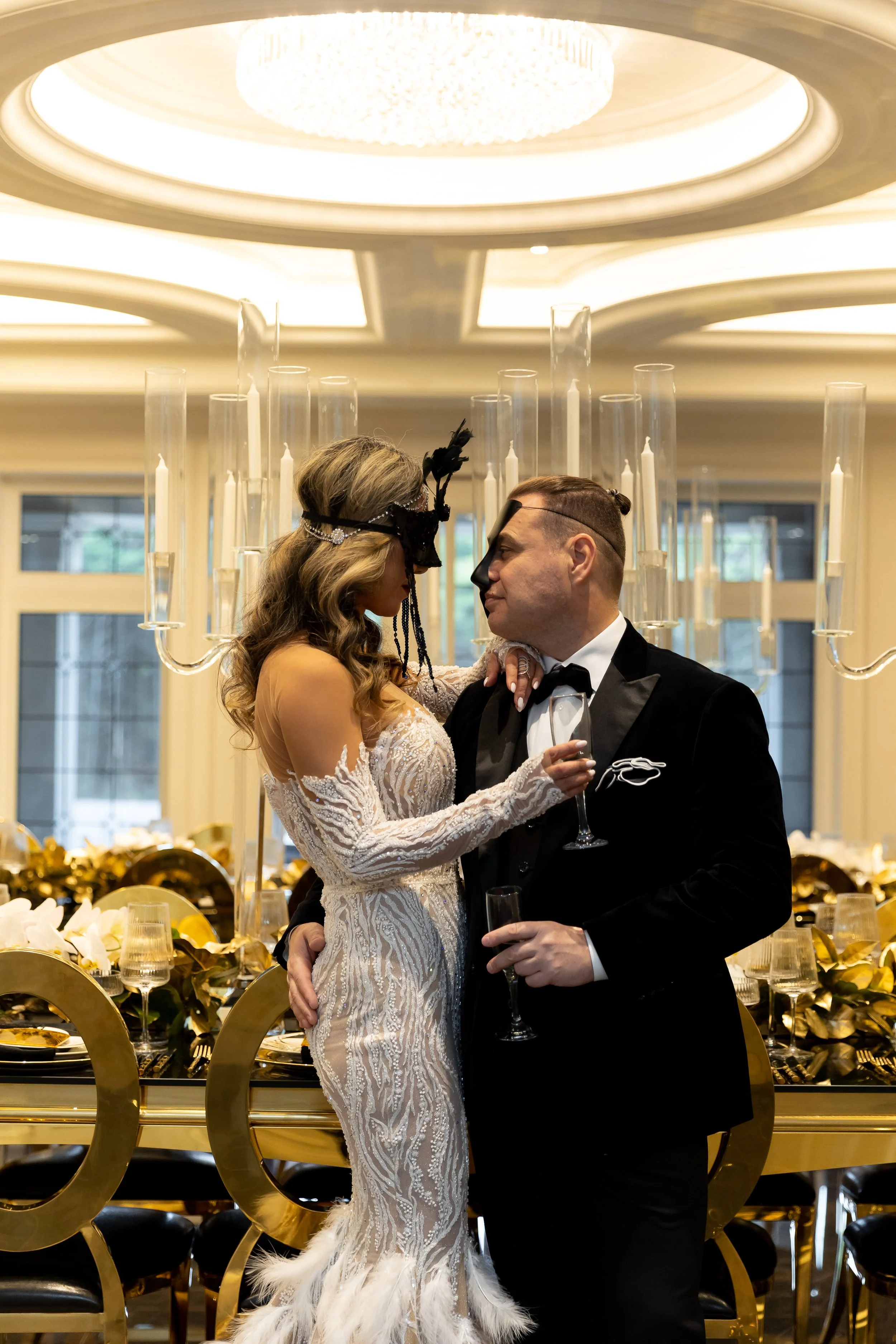 Couple at a wedding reception, decorated with gold and black accents, holding glasses of champagne, with a chandelier overhead and a decorated dining table behind them.