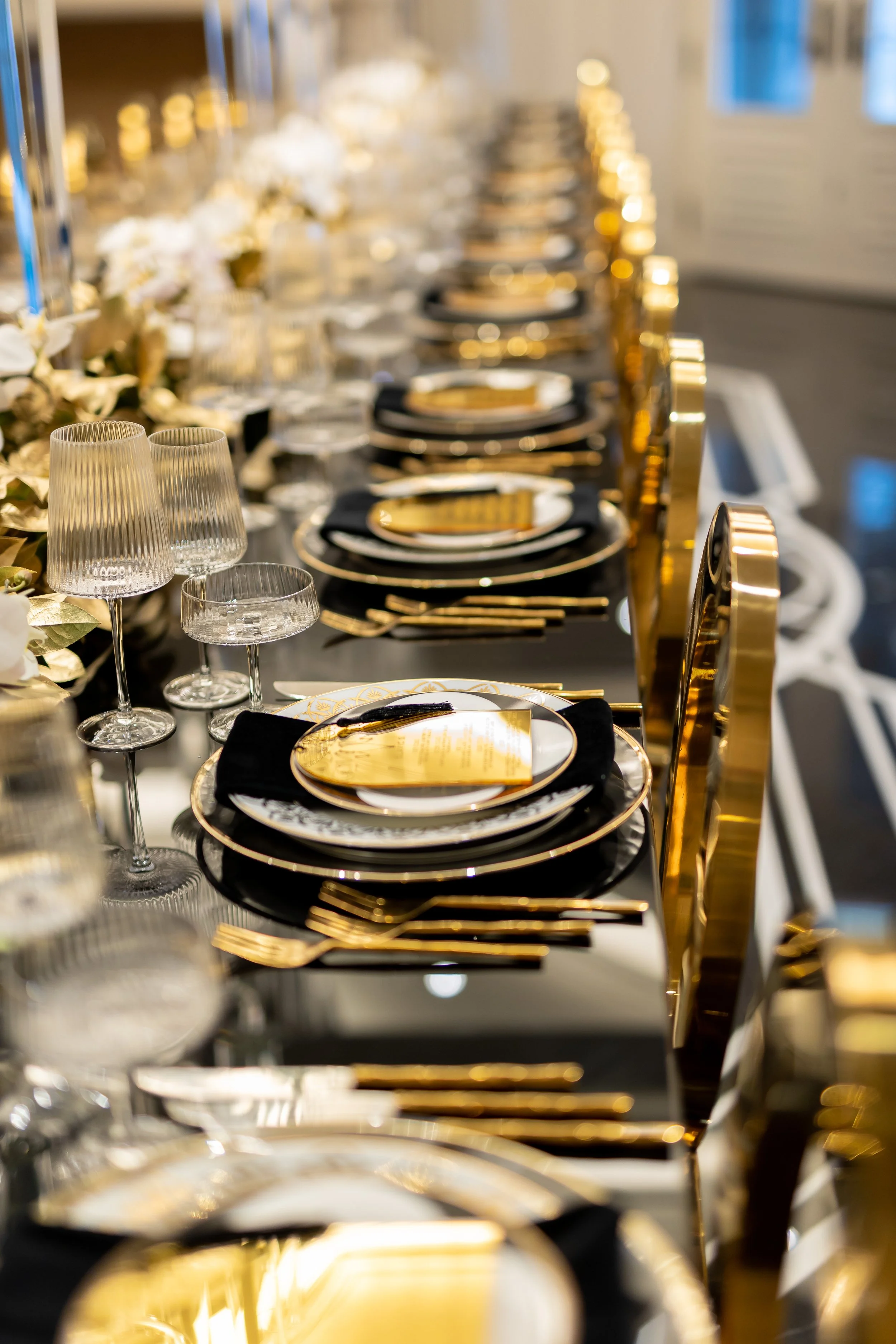 Elegant dining table with black and gold color scheme, gold-rimmed plates, gold flatware, black napkins, and glassware, set for a formal event.