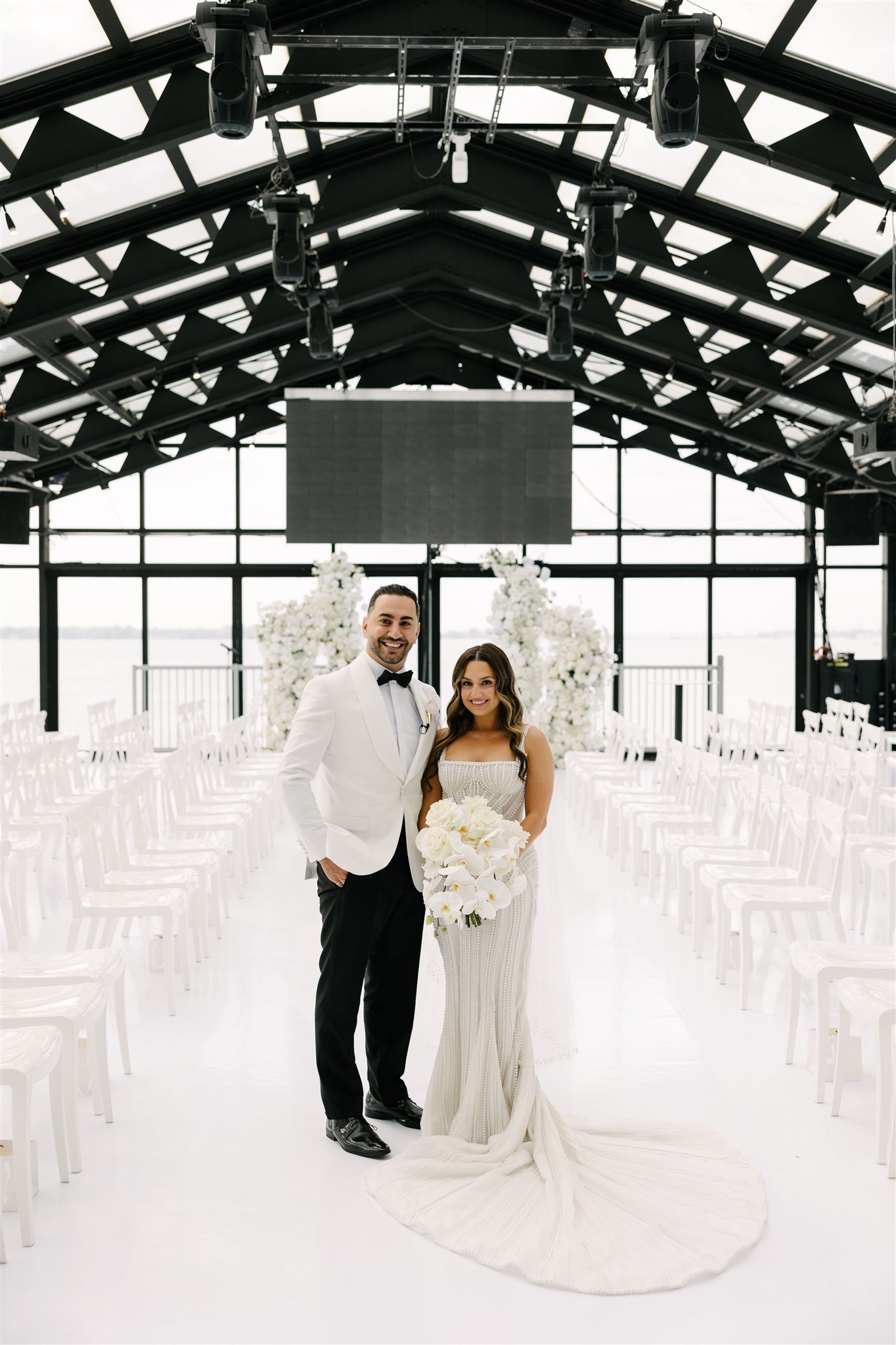A bride and groom standing together in a modern indoor wedding venue with large windows, white chairs, and floral decorations, smiling for the camera.