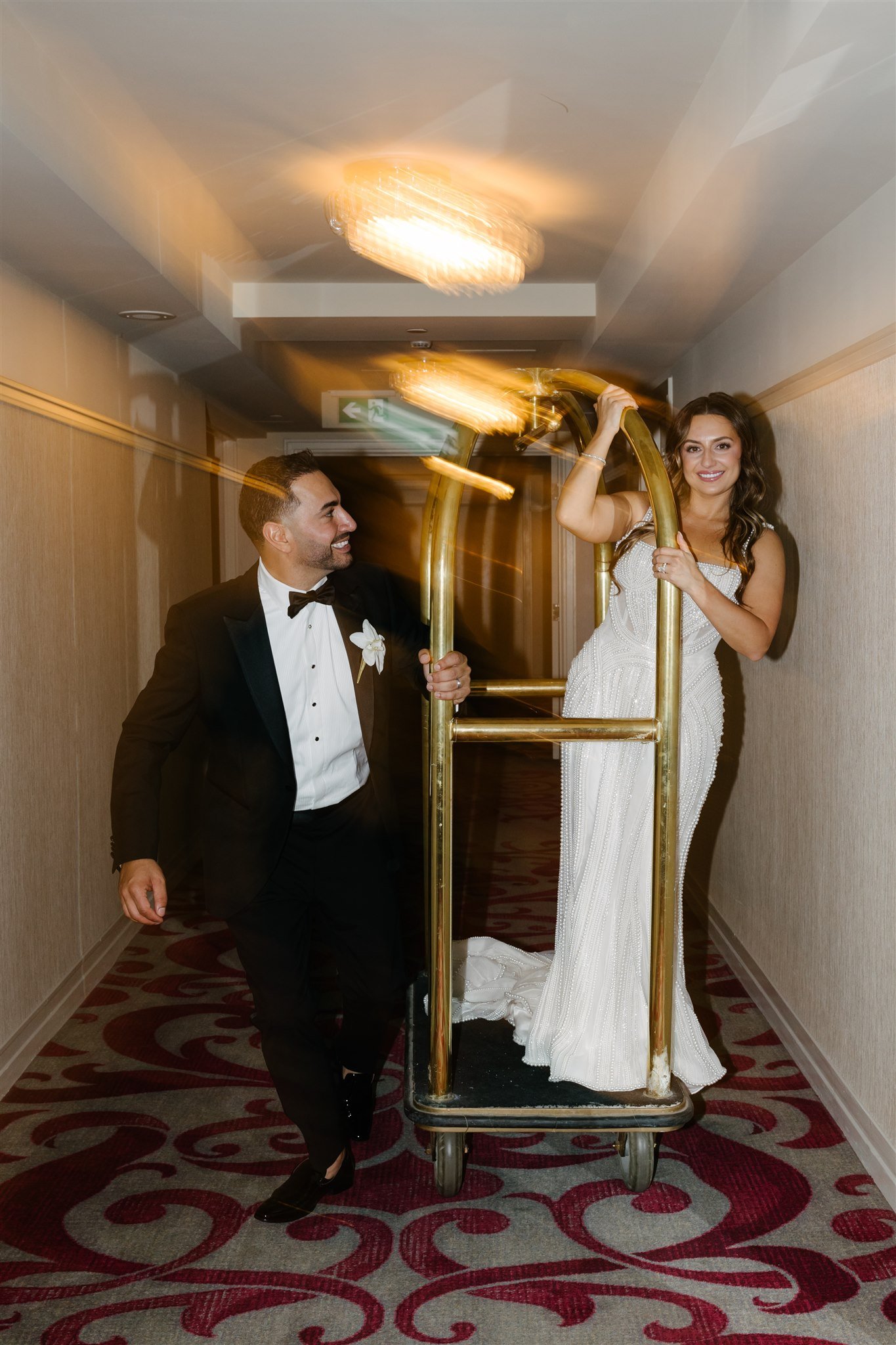 A bride in a white wedding dress and a groom in a black tuxedo with a bow tie riding a luggage cart together in a hotel hallway. The bride is standing on the cart, smiling, while the groom pushes it, both appearing joyful.