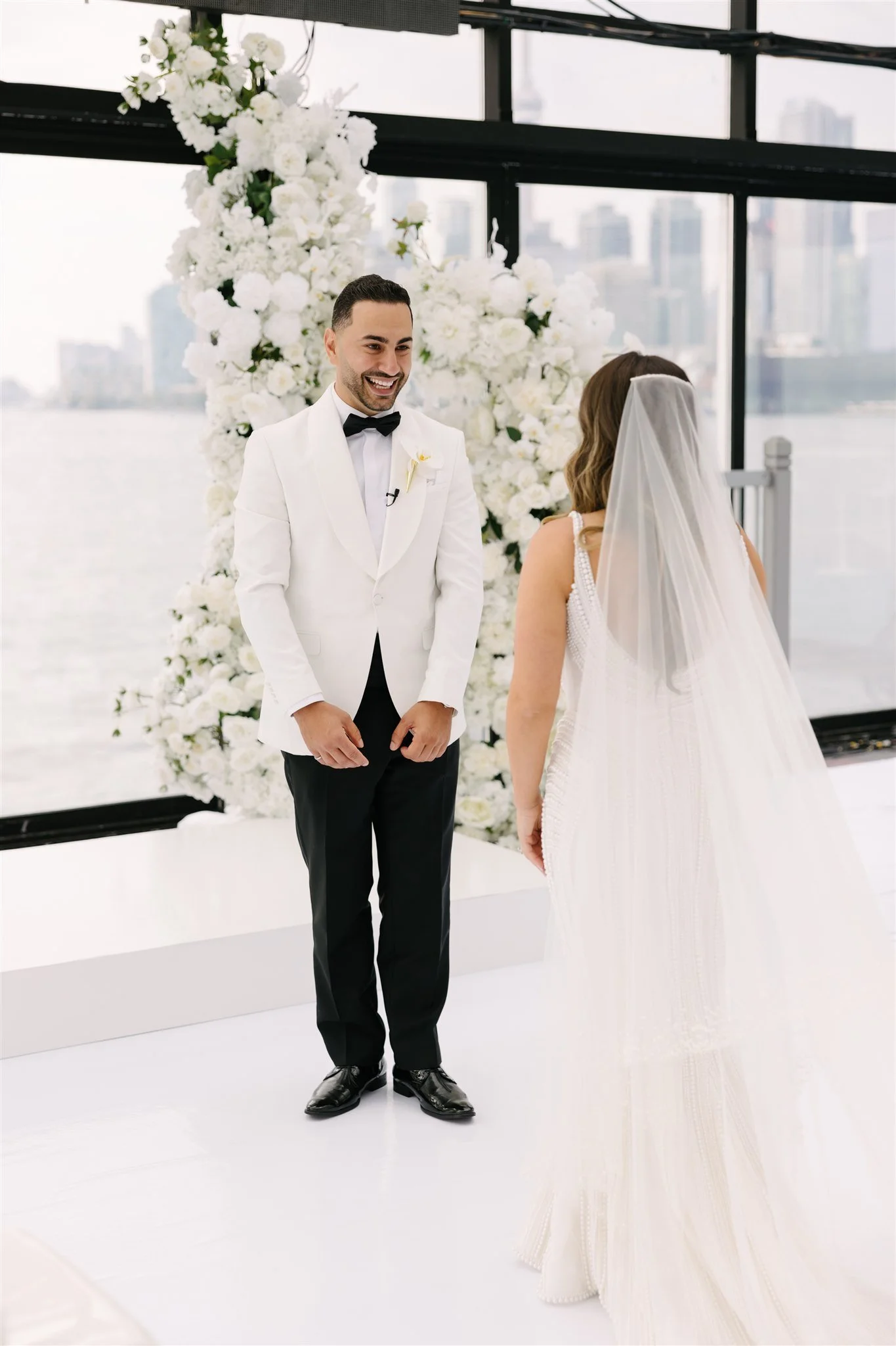 A groom in a white tuxedo jacket and black pants standing opposite a bride in a white dress and veil during a wedding ceremony, with a backdrop of white flowers and large windows overlooking a city skyline and river.