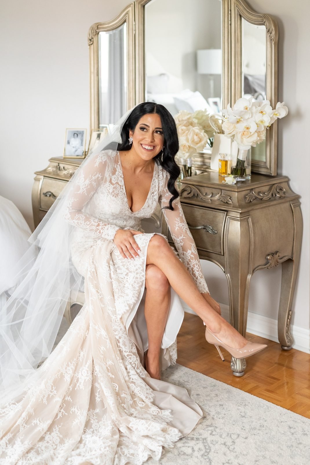 A bride with long dark hair wearing a lace wedding dress and nude heels sitting on the edge of a bed in a bedroom with a vintage dresser and mirror. She is smiling and adjusting her shoe, with a veil draped over her shoulder. A vase with white flower