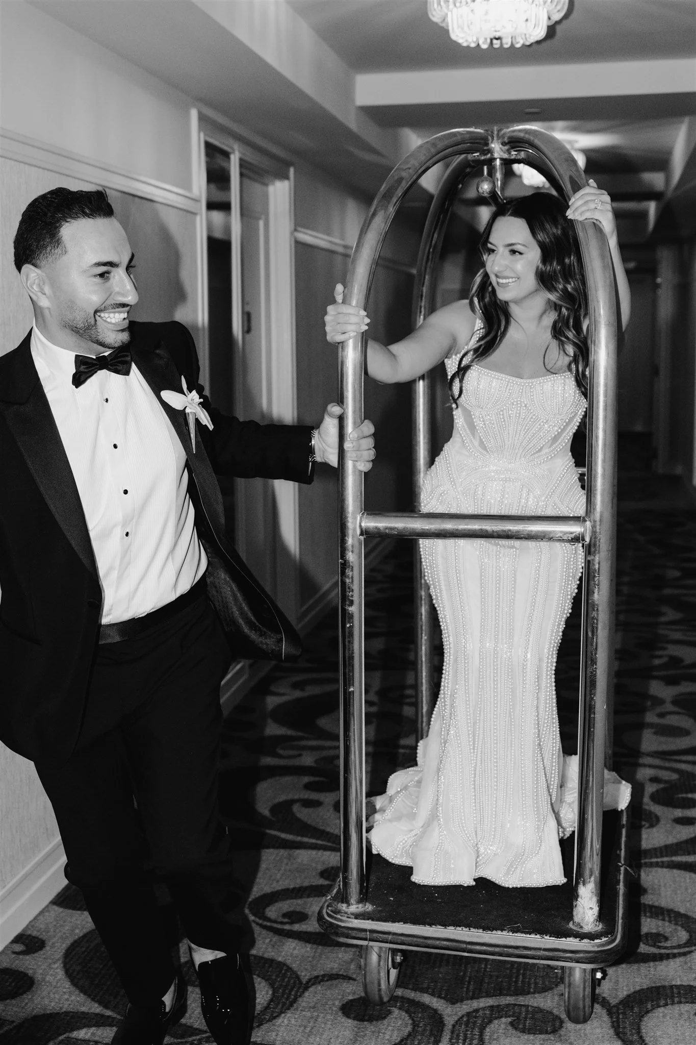 A woman in a wedding dress standing on a luggage cart and a man in a tuxedo pushing the cart, both smiling in a hotel hallway.