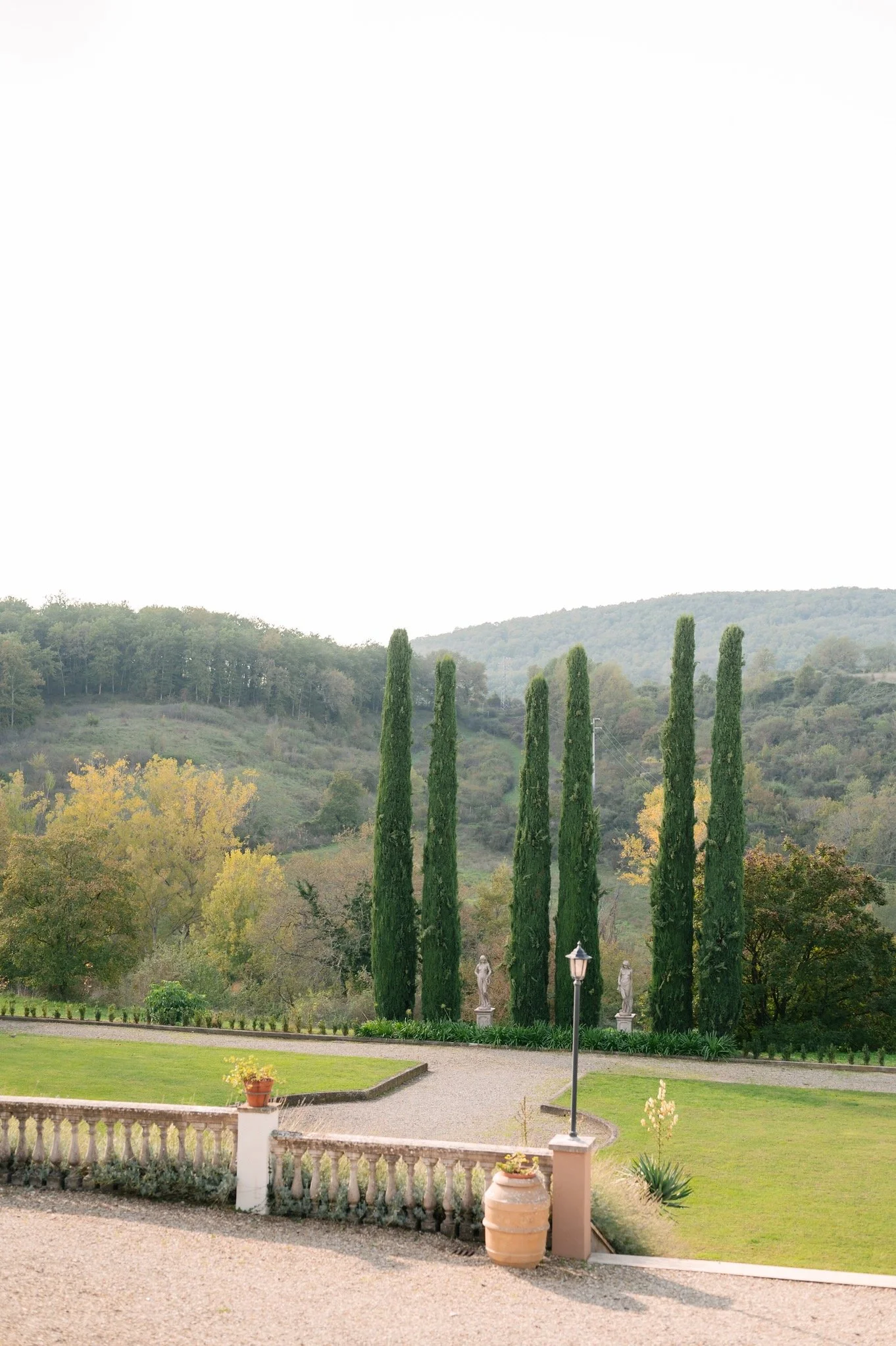 Italian garden with tall cypress trees, ornamental statues, a lamp post, and terracotta pots, set against rolling hills with scattered trees.