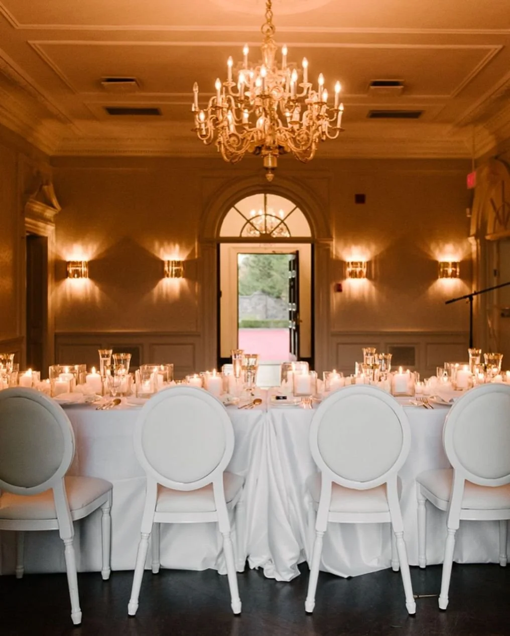 Elegant banquet room decorated for an event with a large chandelier, wall sconces, and a long table set with candles, glassware, and white chairs.