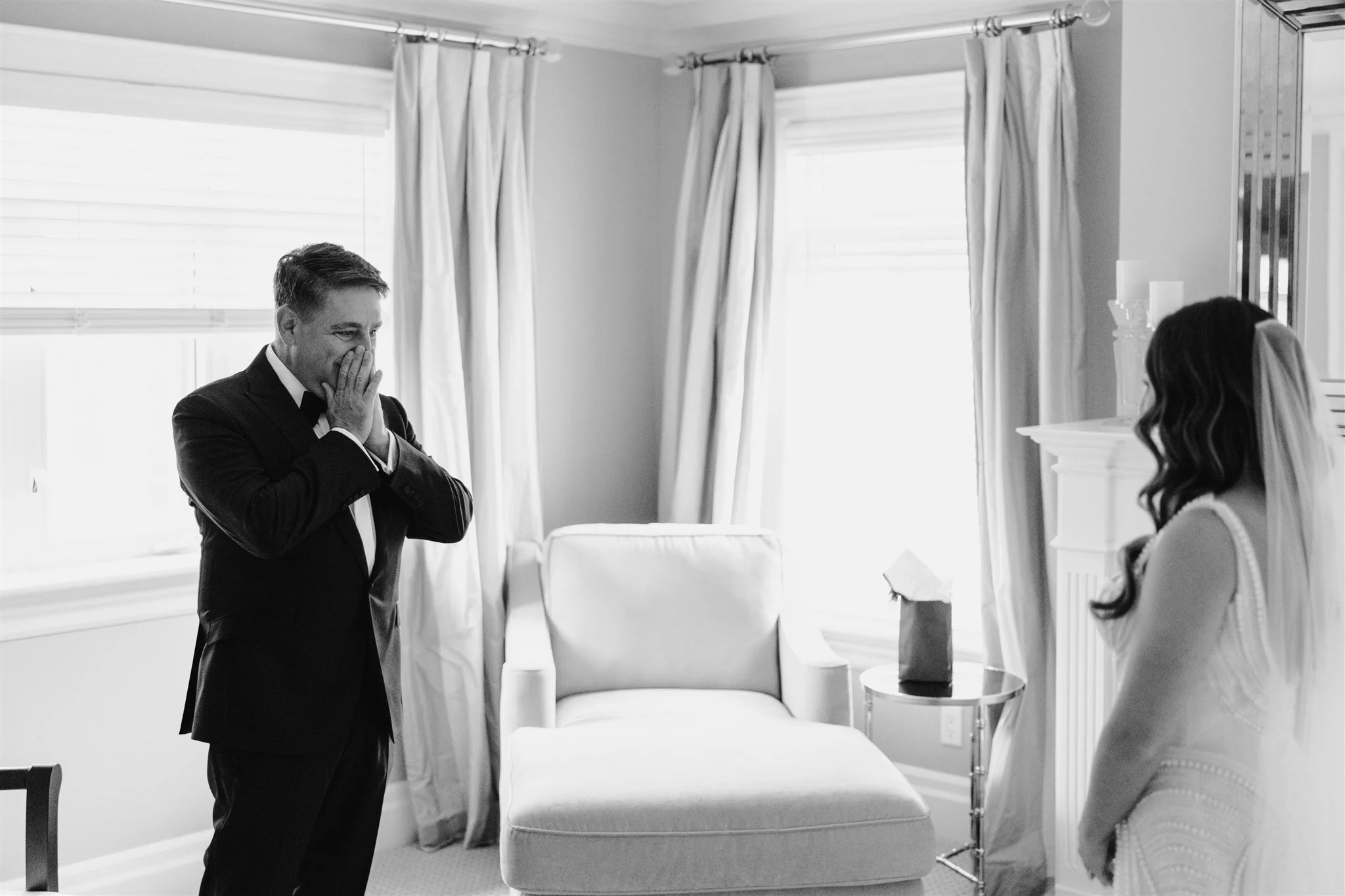 A man in a suit with a bow tie, covering his face with both hands, appears emotional. A woman in a wedding dress and veil is facing him, looking at him. They are in a bright room with windows, curtains, and a chair between them.