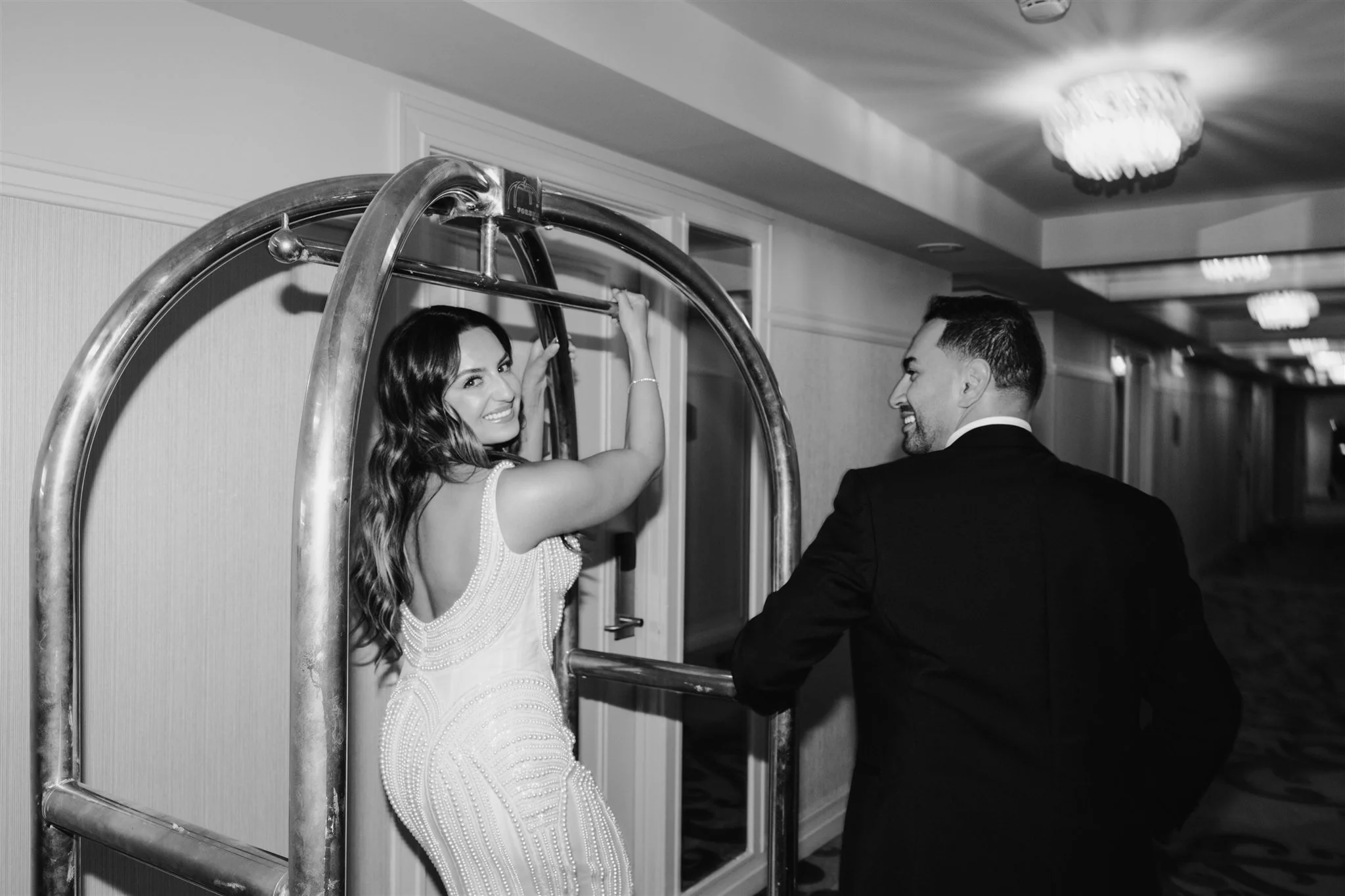 A woman in a glamorous gown and a man in a tuxedo are smiling at each other in a hotel hallway. The woman is standing on a luggage cart.