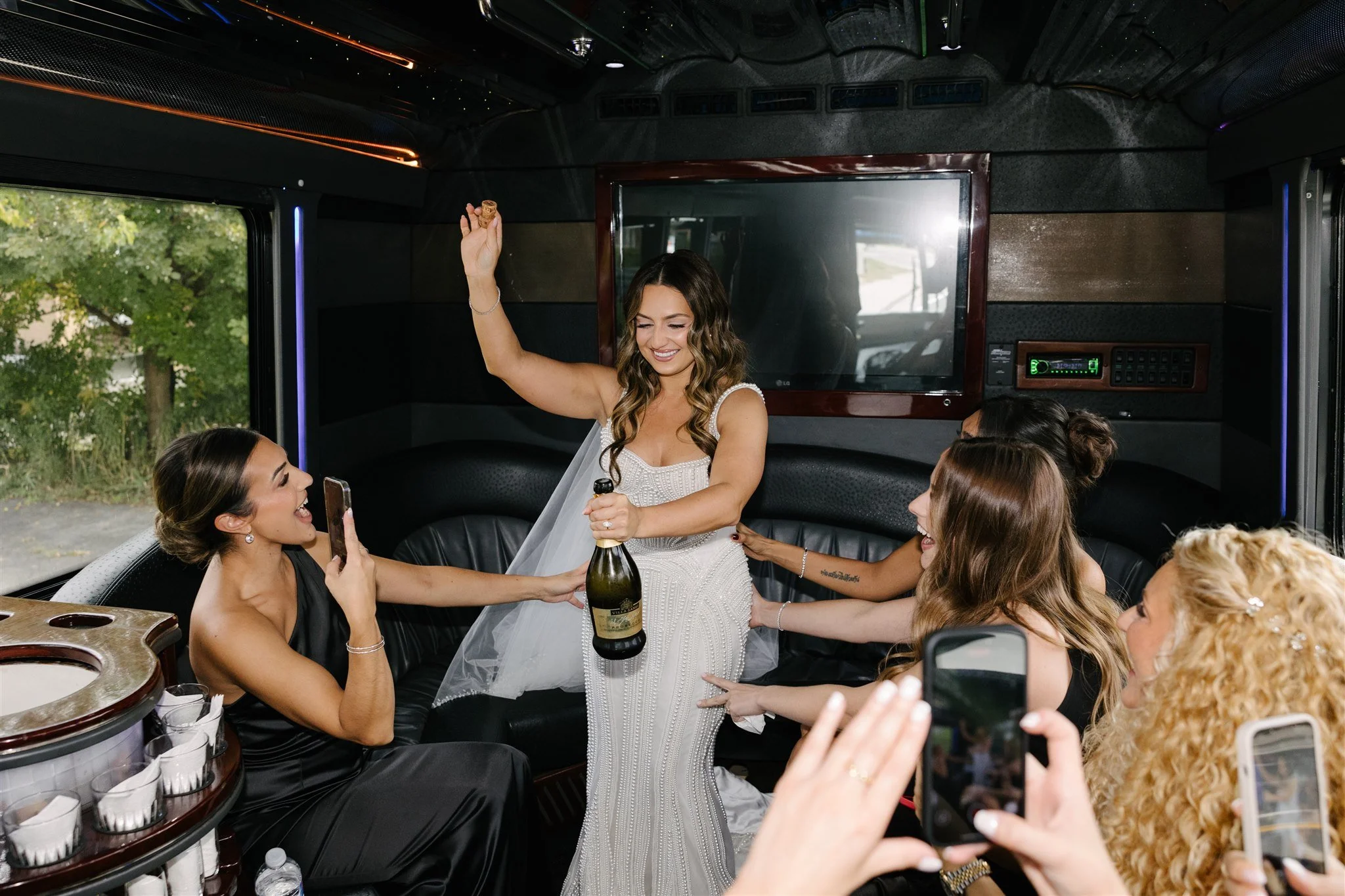 A young woman in a white wedding dress celebrating with friends inside a limousine, holding a bottle of champagne, as friends take photos and cheer.