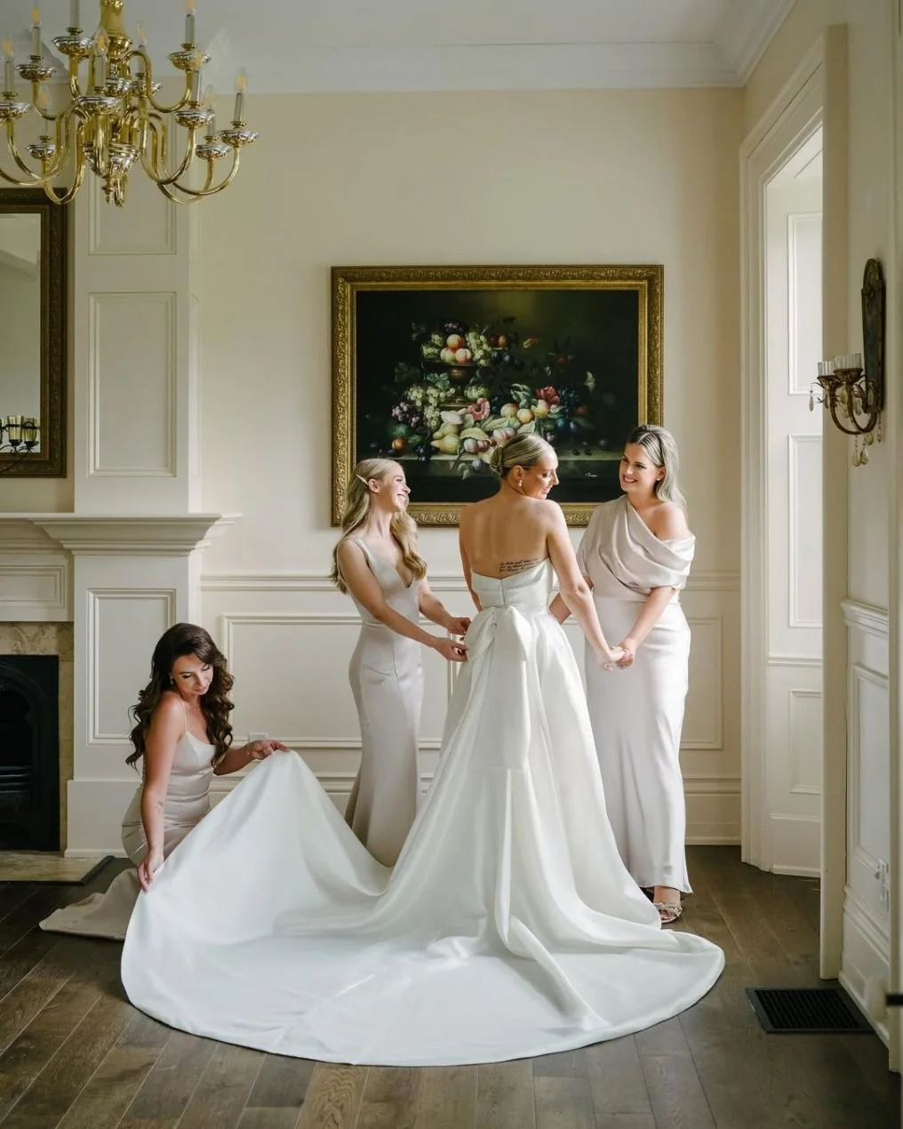 A bride in a white wedding gown is getting ready with her bridesmaids in a bright, elegant room. The bride is surrounded by three women helping her, two of whom are holding her hands and another adjusting her dress. The room features a chandelier, a 