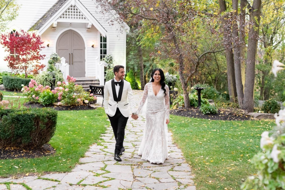 A bride and groom walking hand in hand on a stone path in a garden with trees and colorful flowers, near a white church or house in the background.