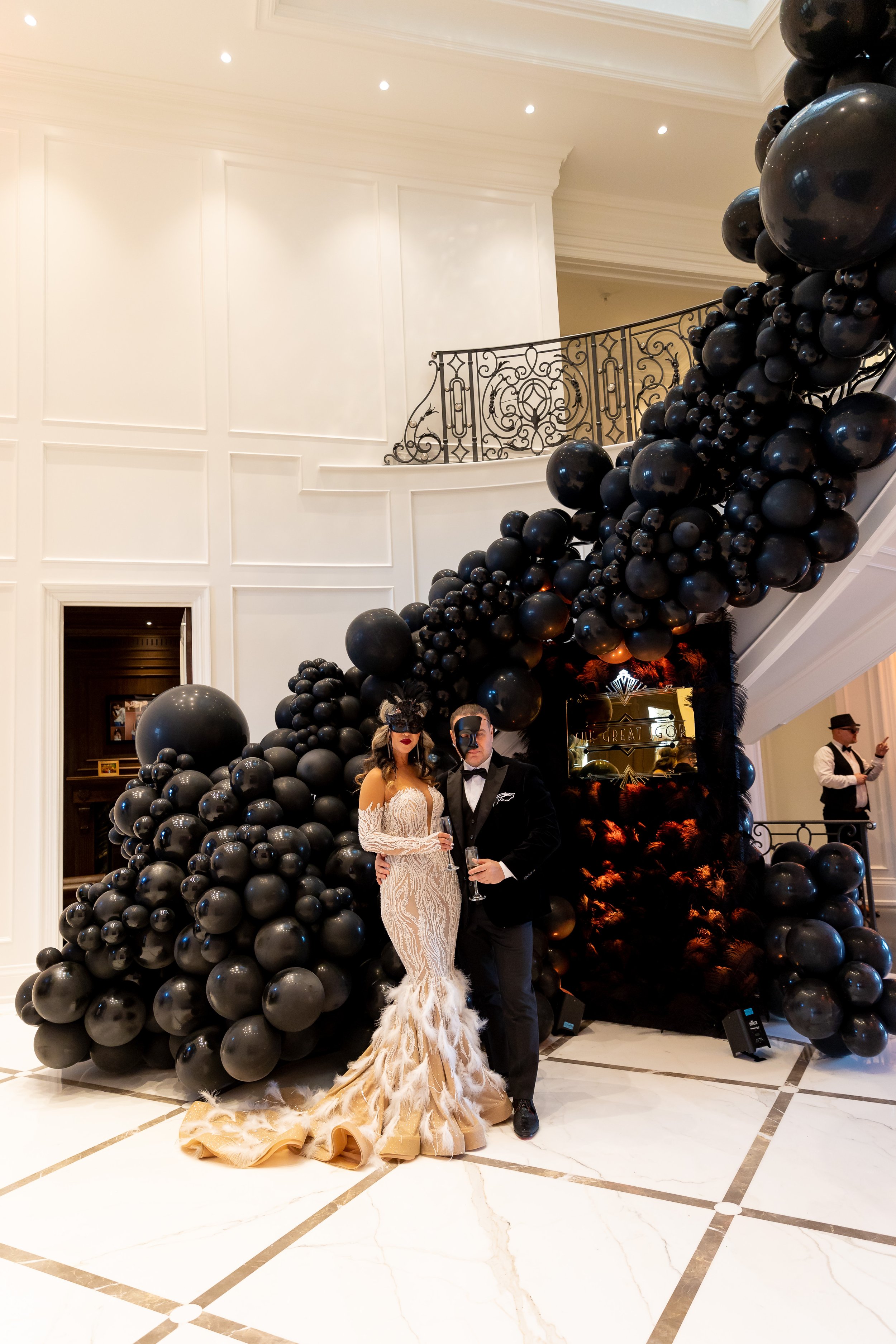 A couple dressed in formal attire posing in front of a balloon arch at a fancy event, with a woman in a feathered gown and a man in a tuxedo, both wearing masks.
