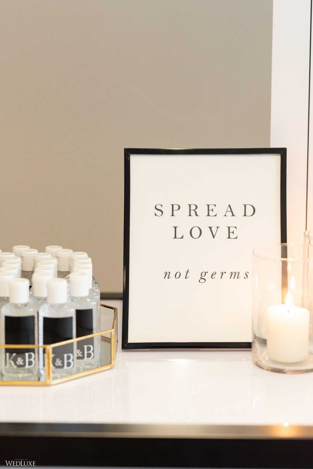 A framed sign reading "Spread Love, not germs" is placed on a table with bottles of hand sanitizer and a lit candle in a glass holder.