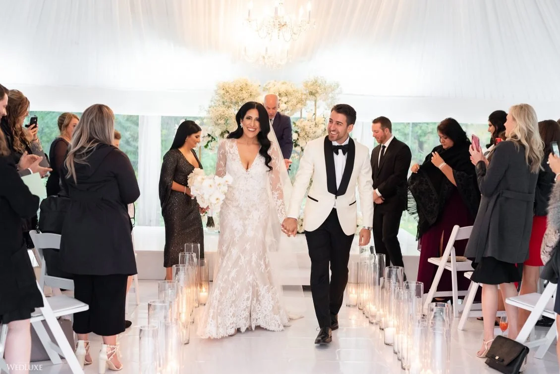 A newly married couple walking down the aisle hand-in-hand in a wedding ceremony, surrounded by seated wedding guests taking photos.