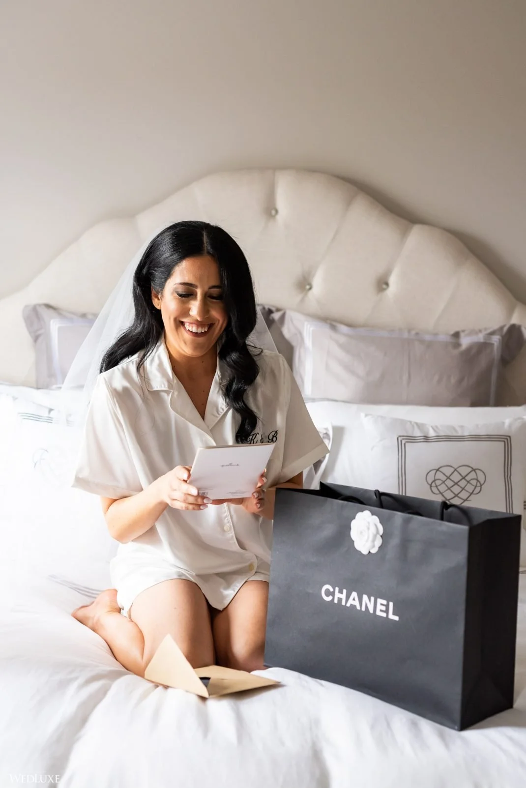 A woman in white pajamas kneels on a bed, opening a gift from Chanel, with a happy expression, in a bedroom with white bedding and pillows.