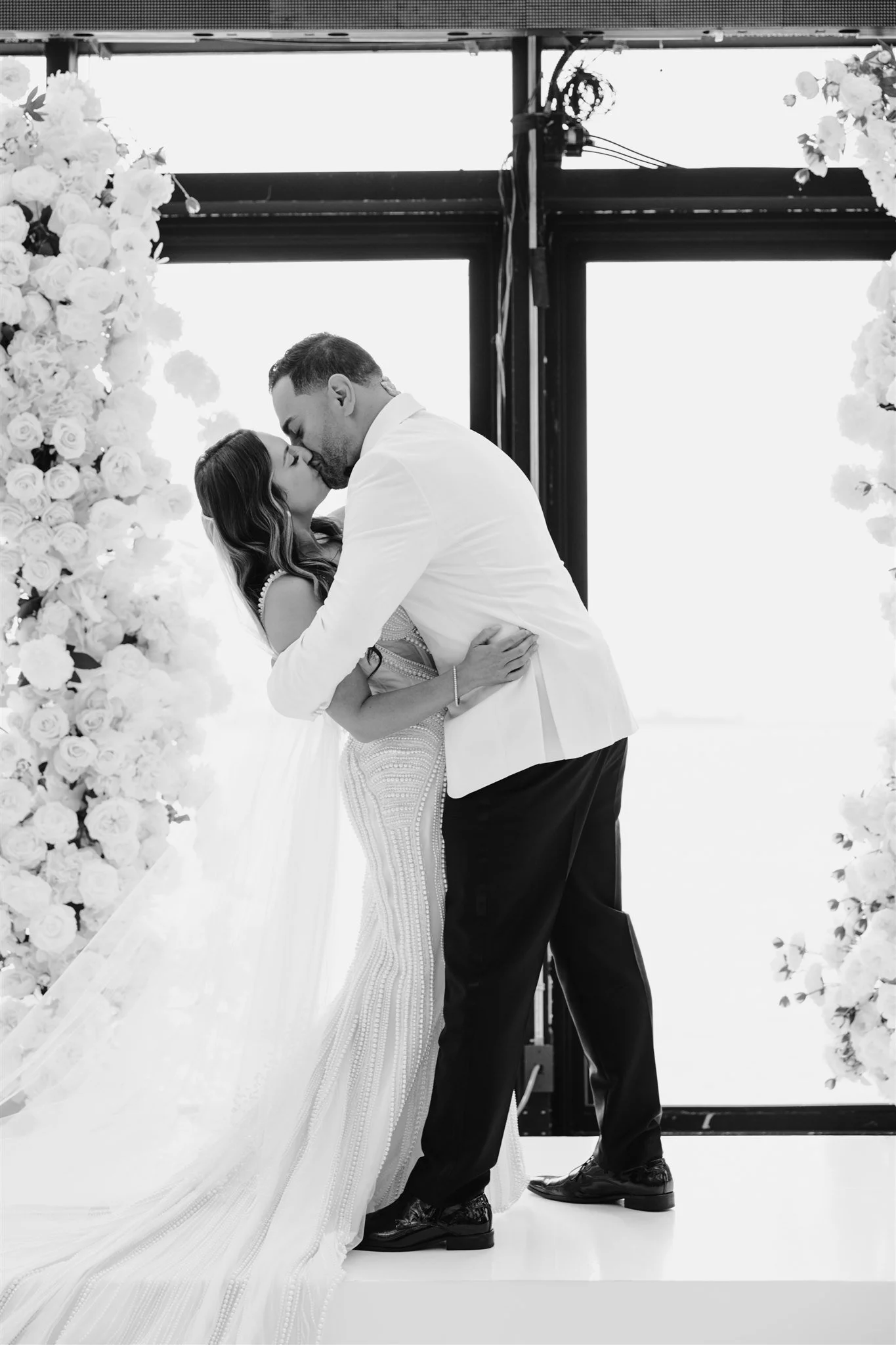 A black and white photo of a bride and groom sharing a kiss during their wedding ceremony, standing on a stage decorated with flowers.