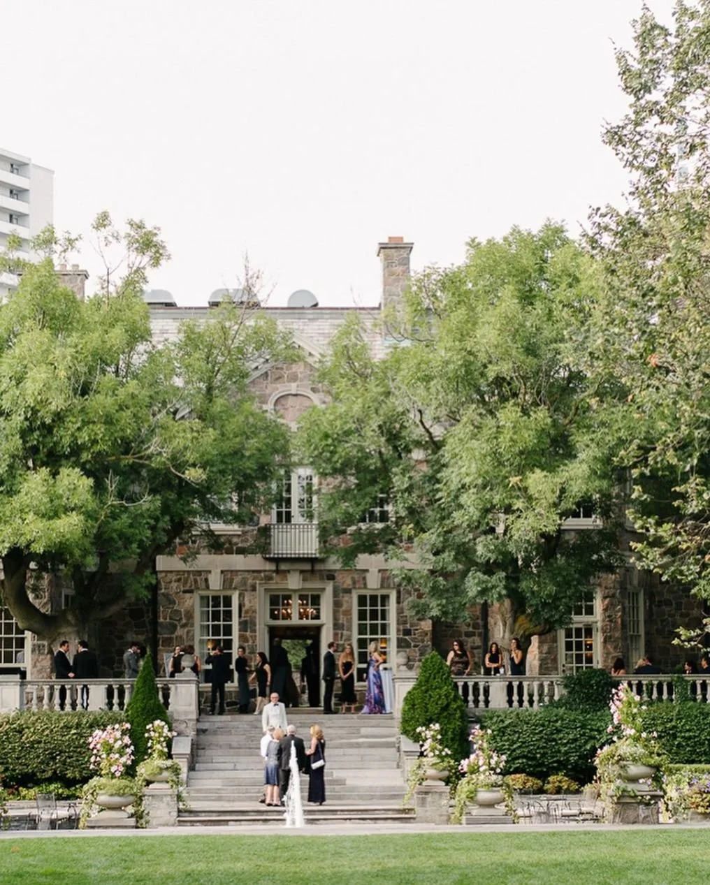 People dressed in formal attire gathering on the steps and in front of a large stone mansion surrounded by trees and bushes for an outdoor event, possibly a wedding.