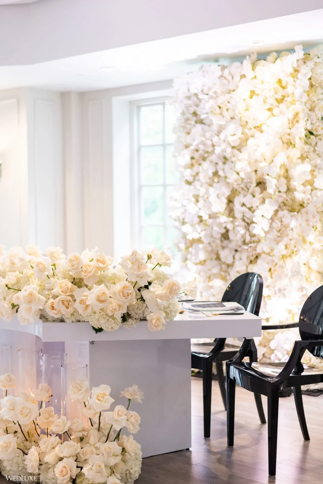 Elegant event space decorated with white roses and hydrangeas, featuring a floral backdrop, black chairs, and a white table adorned with a floral arrangement.