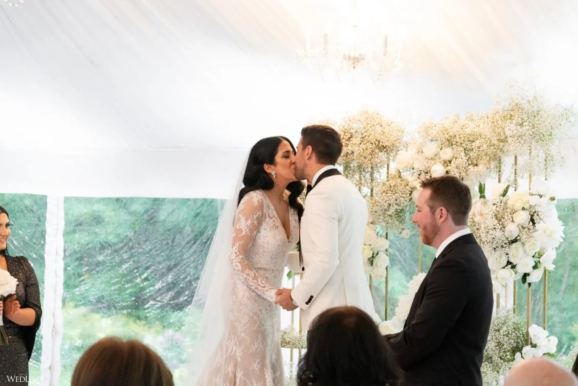 A bride and groom kiss during their wedding ceremony in a decorated tent with white flowers and greenery, surrounded by guests.