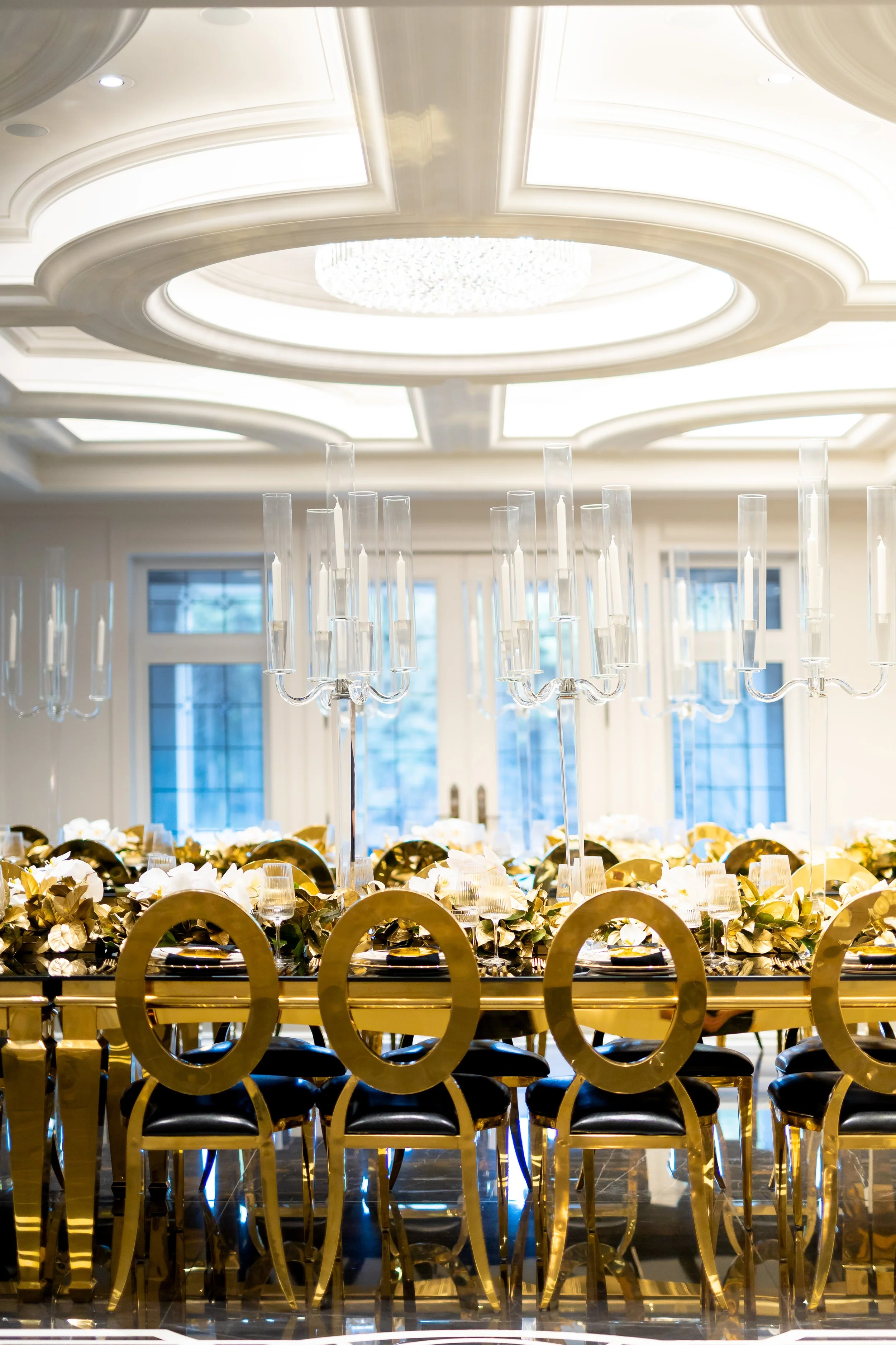 Elegant dining room with a long gold and black table, tall glass candle holders, floral arrangements, black and gold chairs, and a white ornate ceiling with a chandelier.
