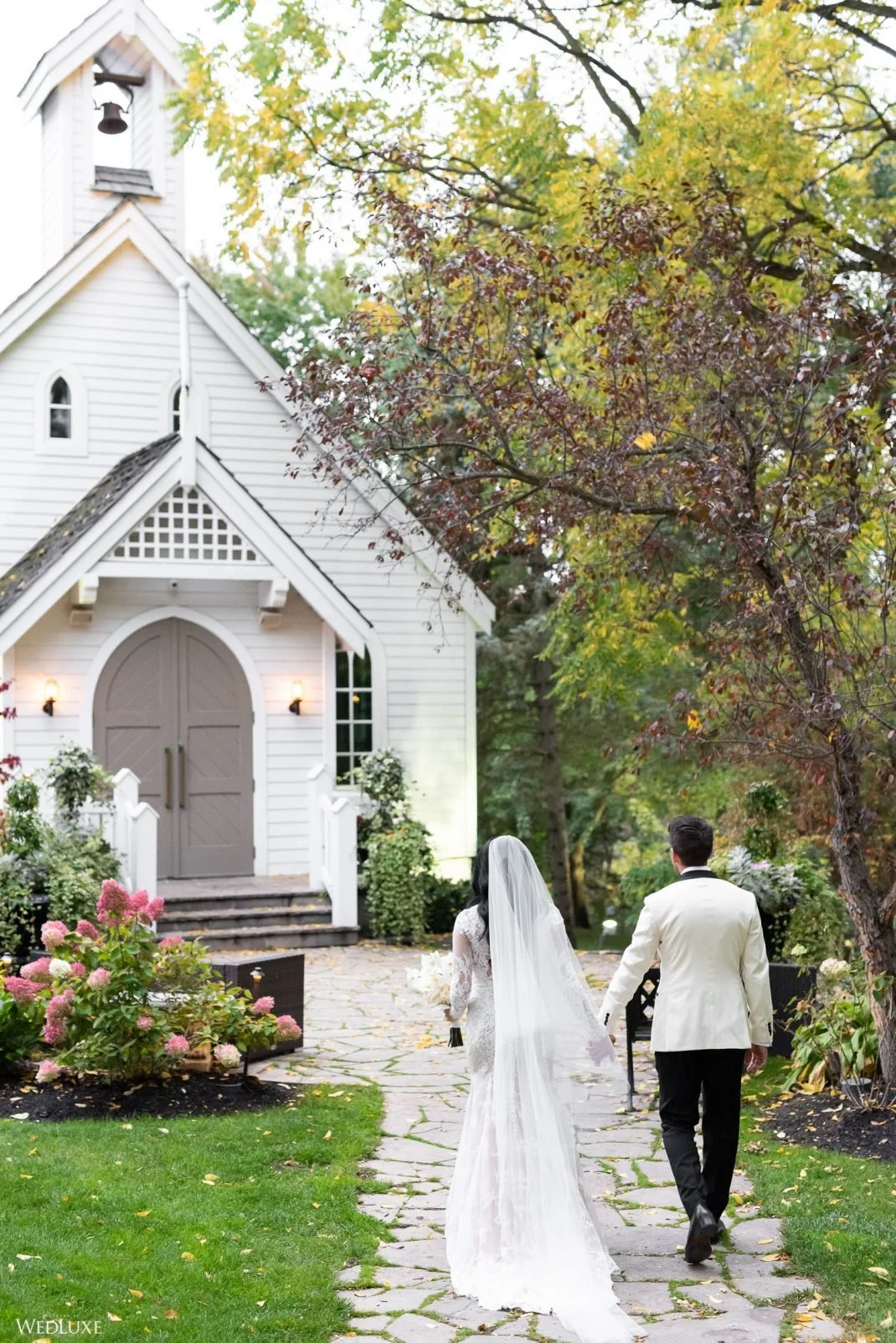 A bride and groom walking hand in hand toward a small white chapel amid colorful autumn trees and flowers.