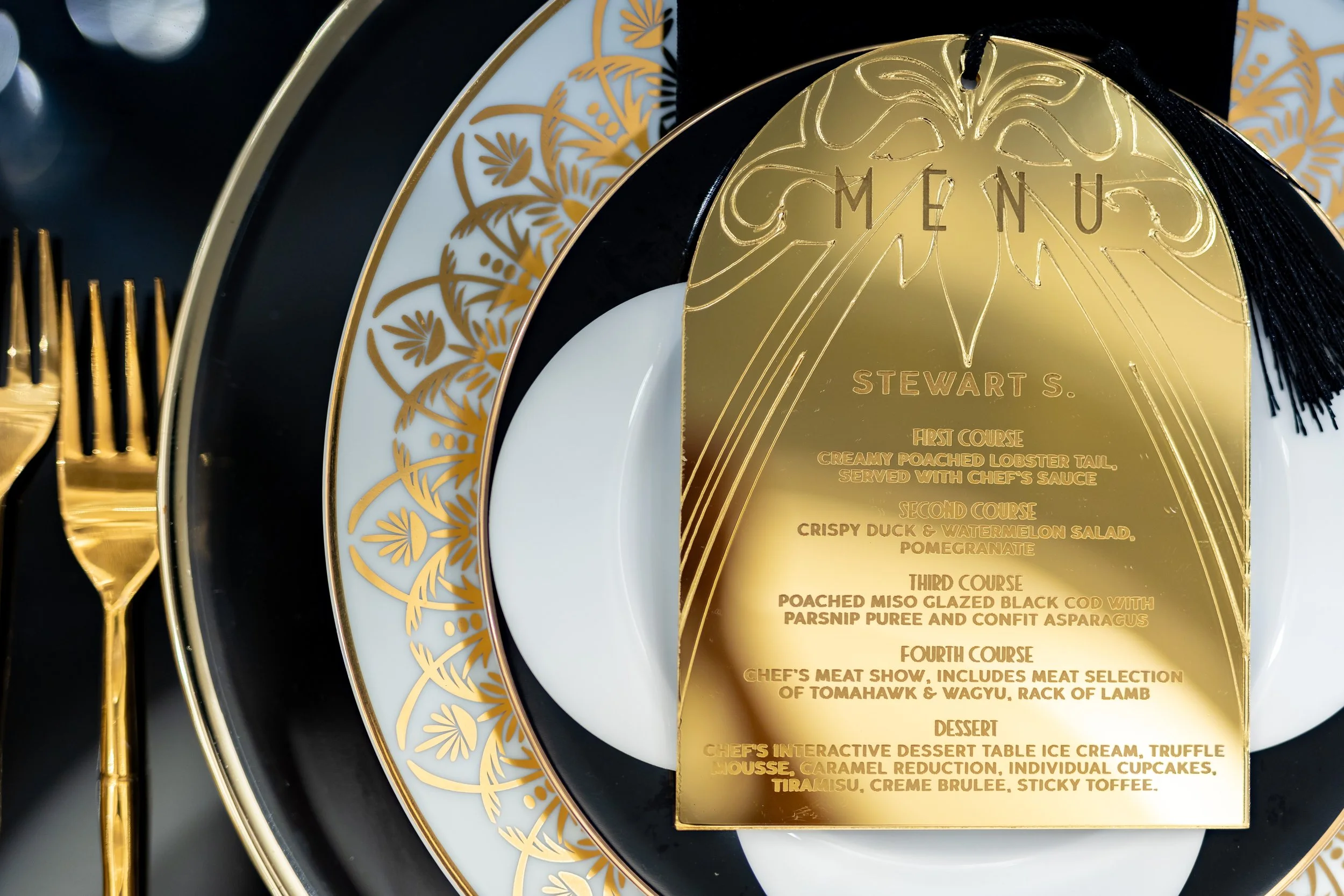 A gold menu card with a decorative pattern at the top, placed on a set of white and black plates, with gold cutlery visible on the left side.