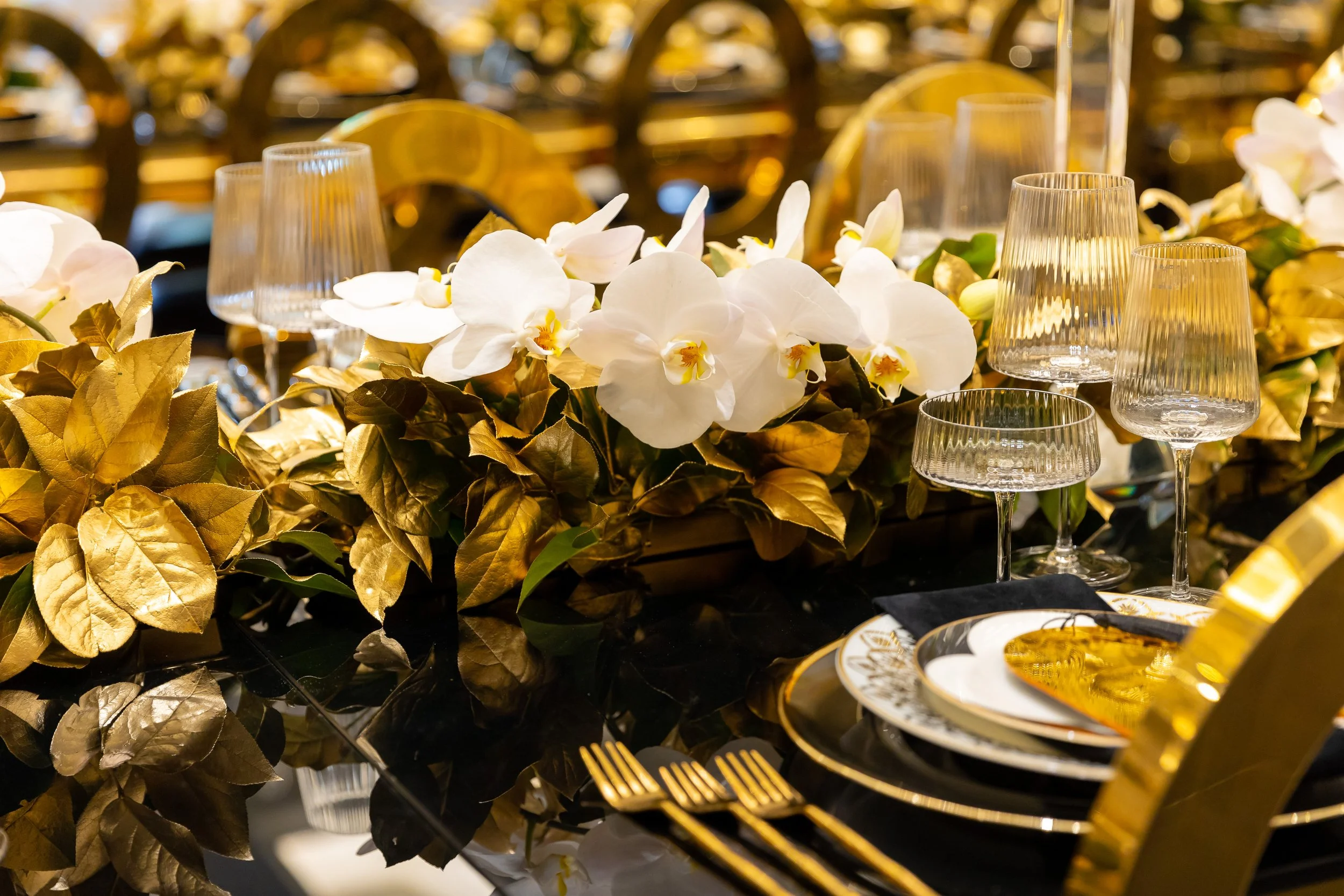 Elegant dining table decorated with white orchids, gold and black tableware, gold cutlery, and gold and clear glassware, with a black reflective table surface.