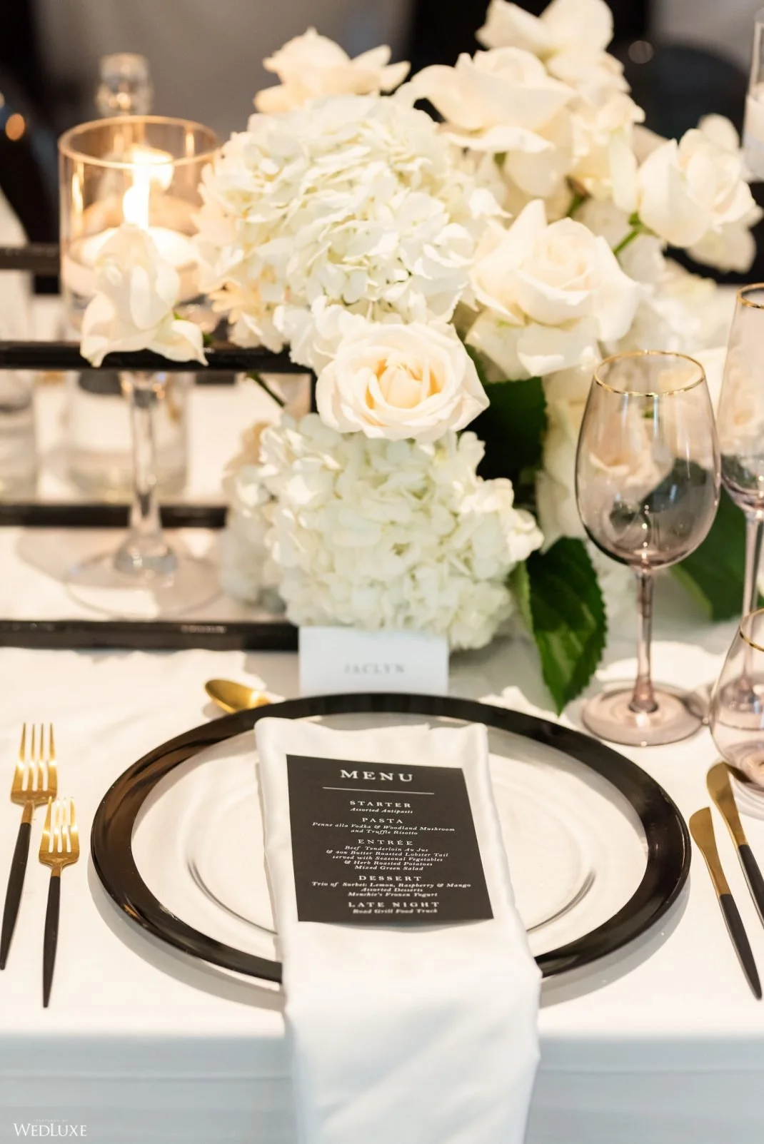 Elegant table setting with a white plate, gold utensils, a black-bordered plate, a white napkin with a menu, a floral centerpiece of white roses and hydrangeas, and a lit candle.