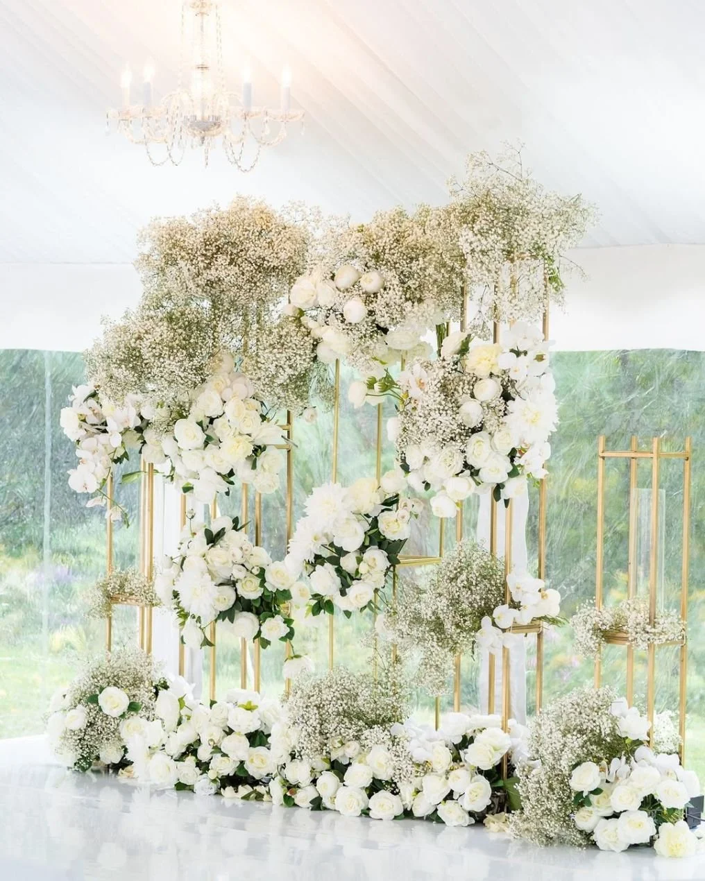 Elegant floral arrangement with white roses, baby's breath, and greenery, set against a backdrop of gold frames and a white ceiling with a chandelier.