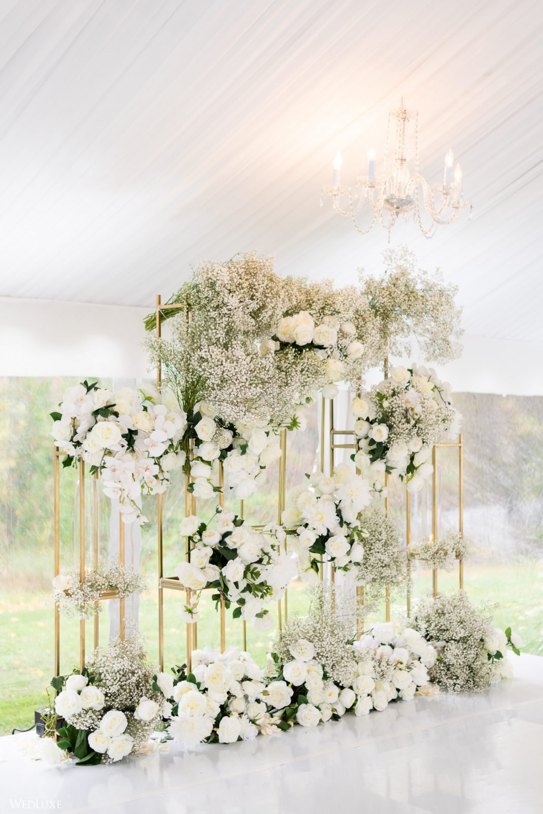 Wedding floral arrangement with white roses, baby's breath, and greenery on gold stands, beneath a chandelier in a bright room with large windows.
