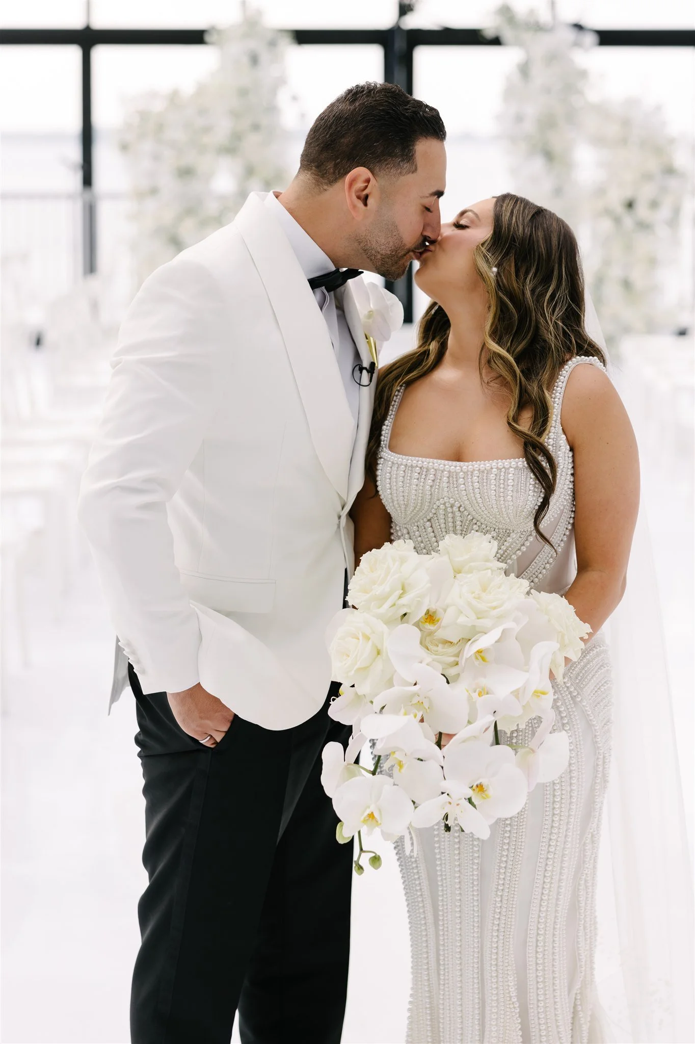 A bride and groom share a kiss at their wedding. The groom is wearing a white tuxedo jacket, black pants, and a bow tie, and the bride is holding a large bouquet of white flowers and wearing a beaded wedding dress.