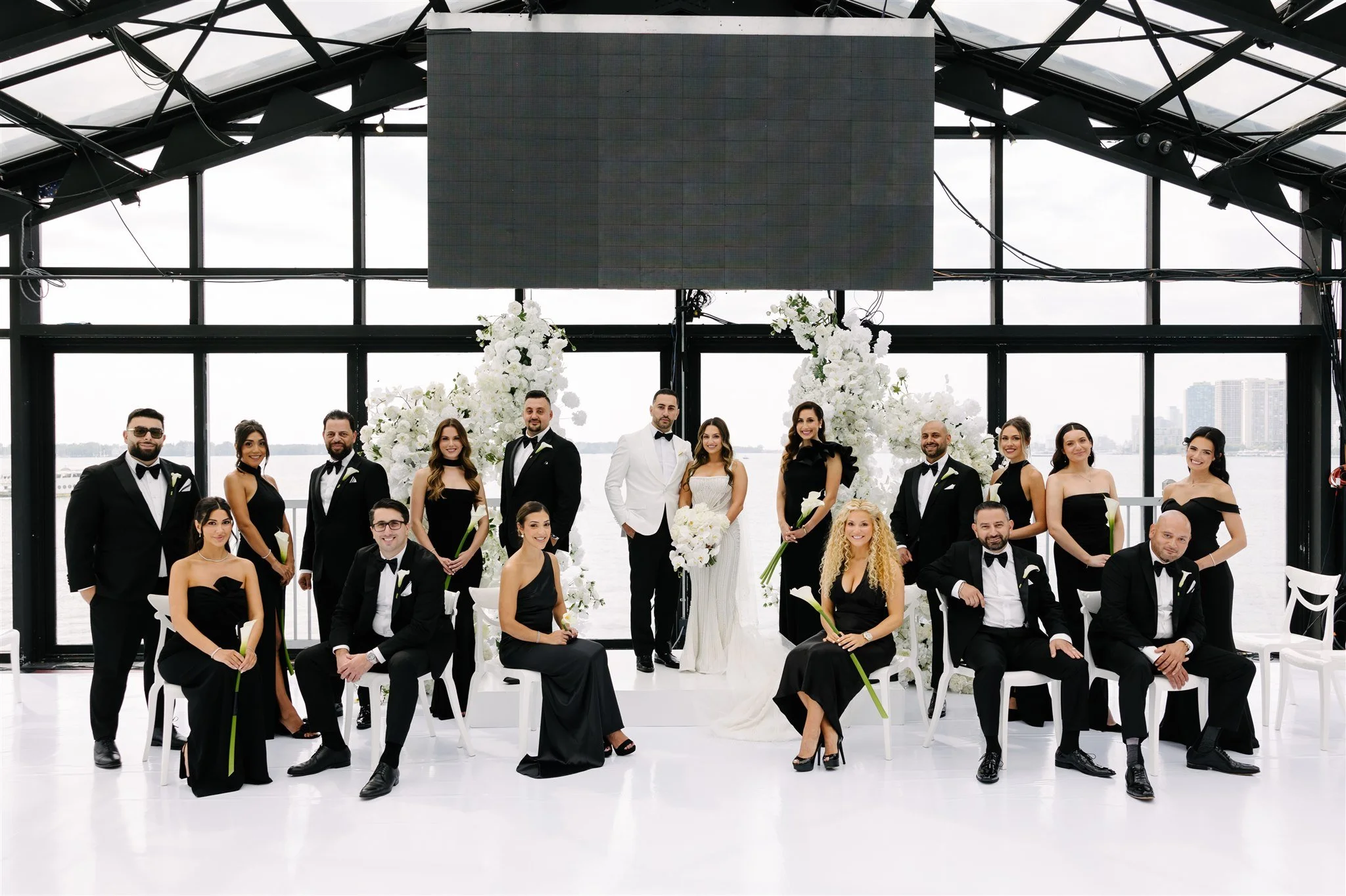 A wedding party with 16 people dressed in formal attire, standing and sitting in front of large windows with a cityscape view and waterfront background. The bride and groom are in the center, surrounded by bridesmaids and groomsmen, with floral decor