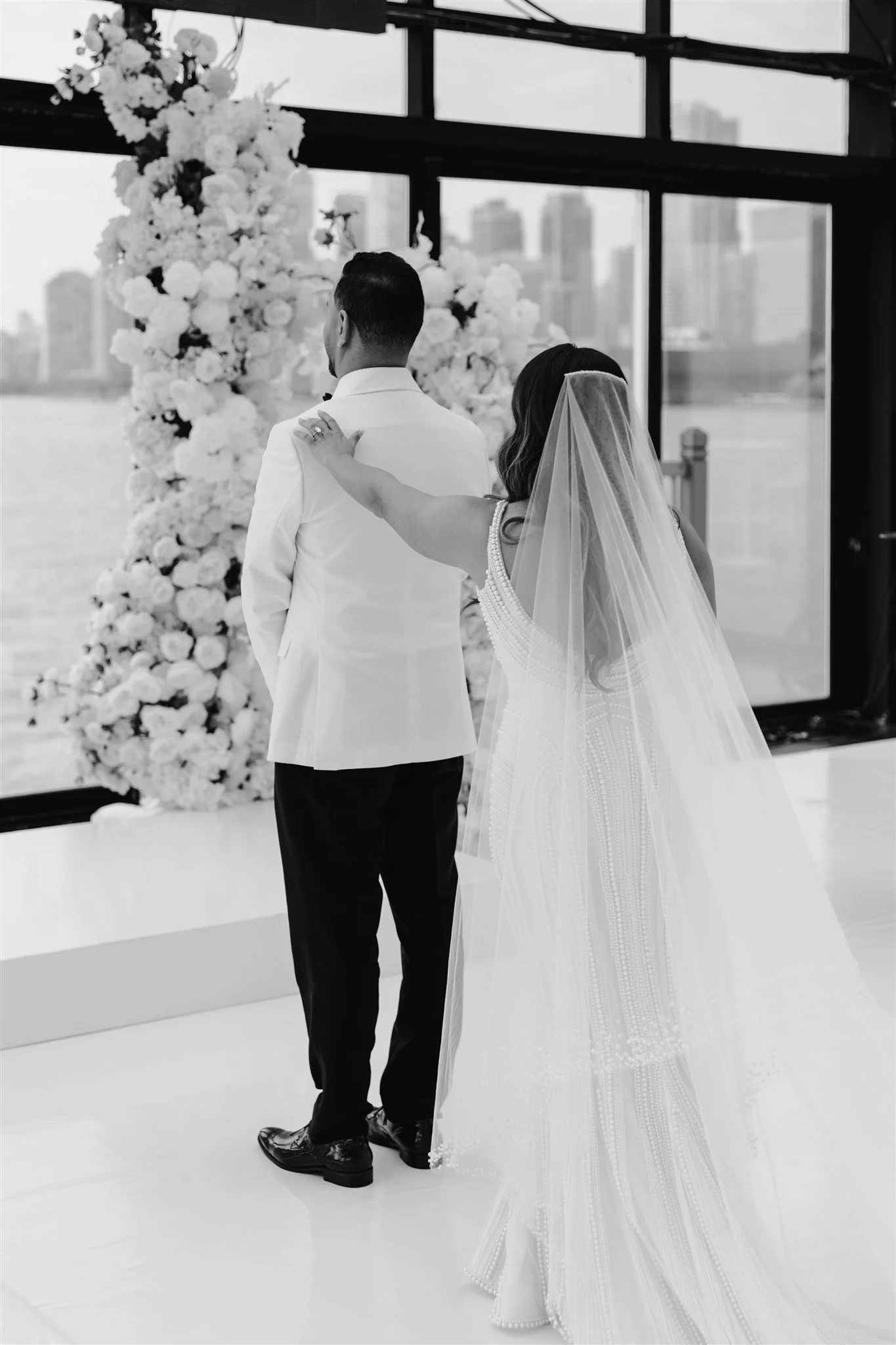 A bride and groom stand together in front of a floral wedding arch, with the bride touching the groom's shoulder, inside a modern building with large windows overlooking a cityscape.
