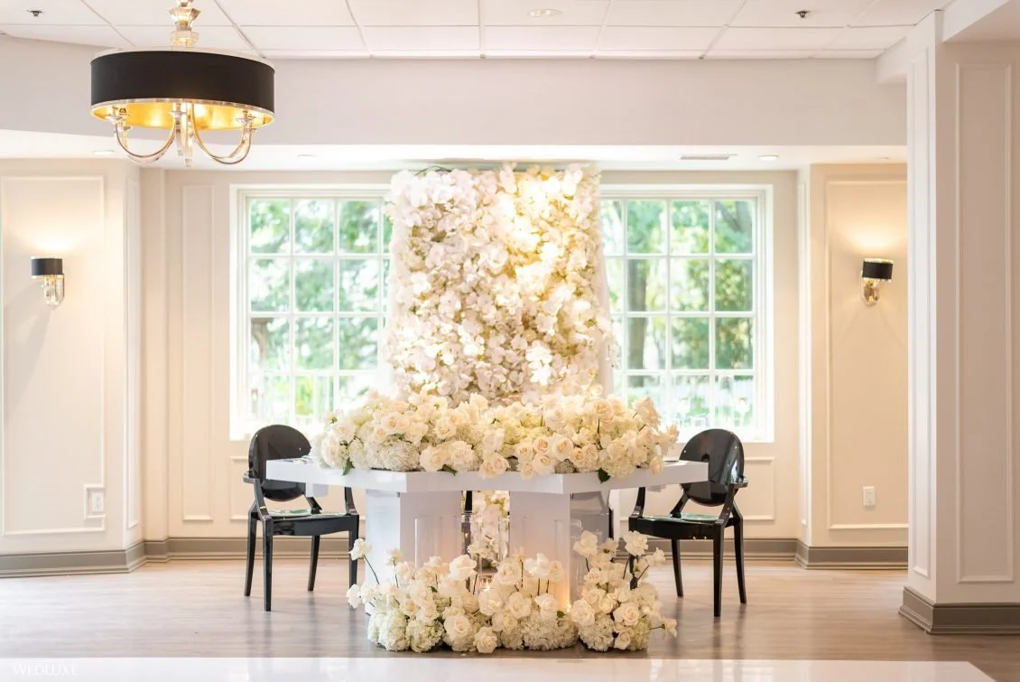 Elegant indoor space with table covered in white flowers and a floral backdrop, two black chairs, large windows showing greenery outside