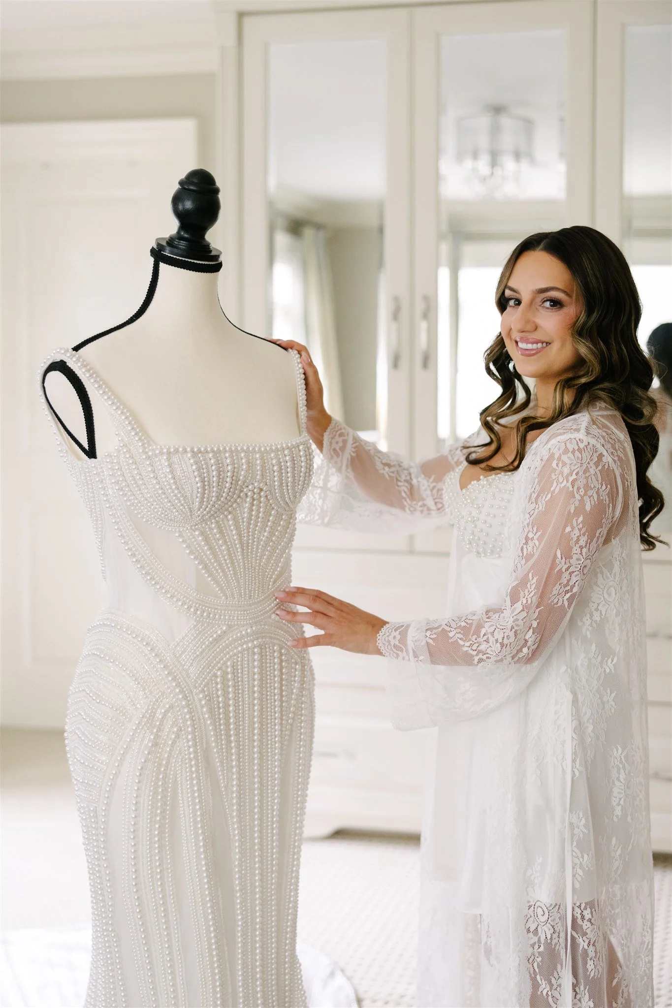 Bride in a lace robe standing next to a mannequin wearing a pearl-embellished wedding dress.