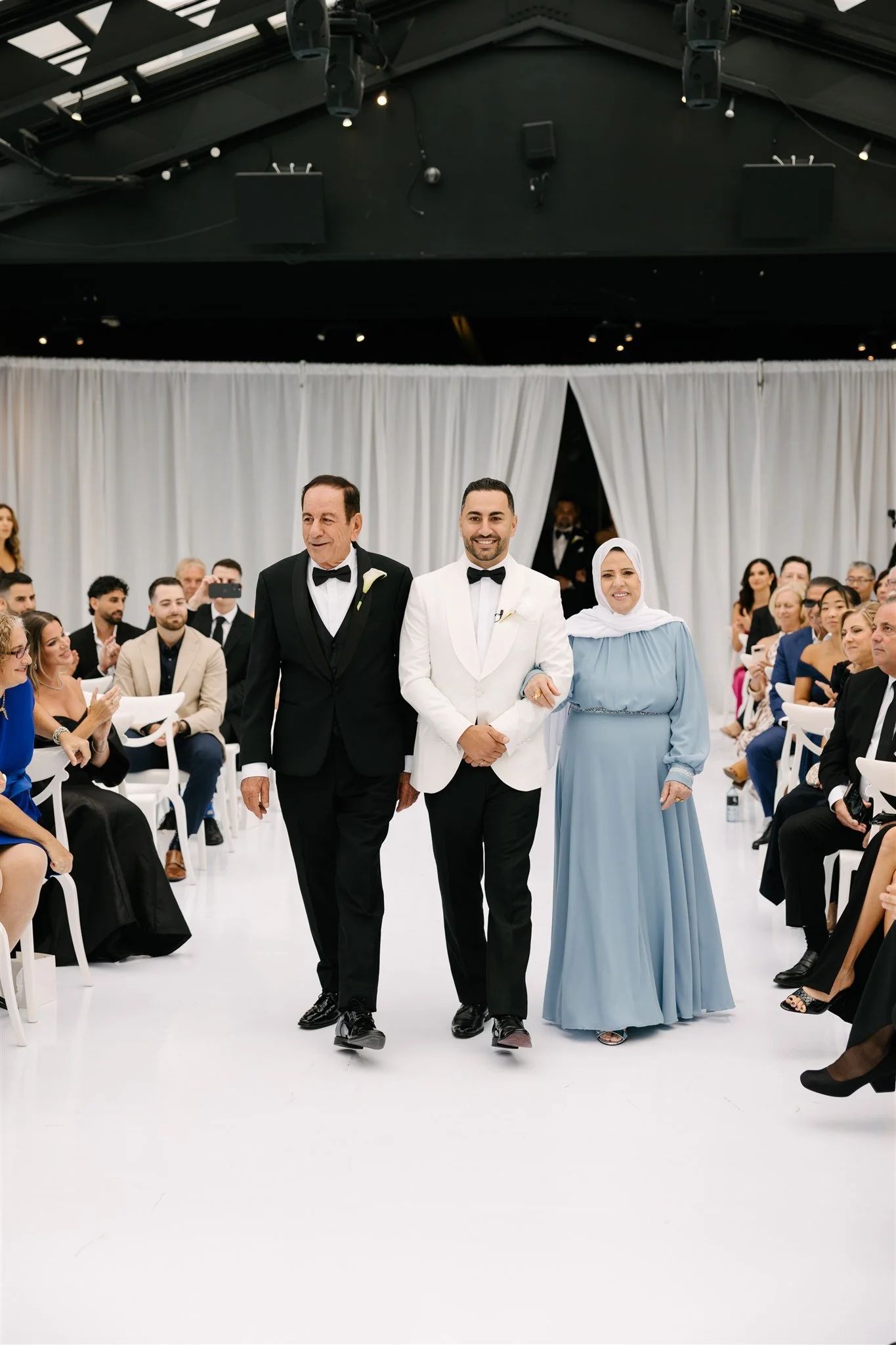 A wedding ceremony with a groom in white tuxedo walking with his parents down the aisle, surrounded by seated guests in an indoor venue with white curtains and black ceiling.