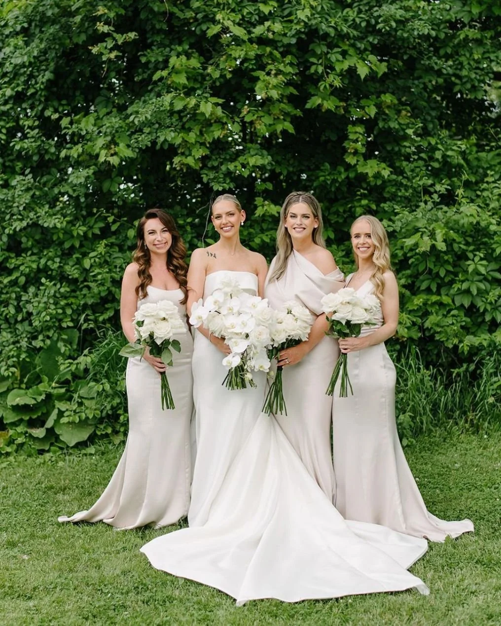Four women in white dresses holding bouquets of white flowers standing outdoors in front of green leafy bushes.