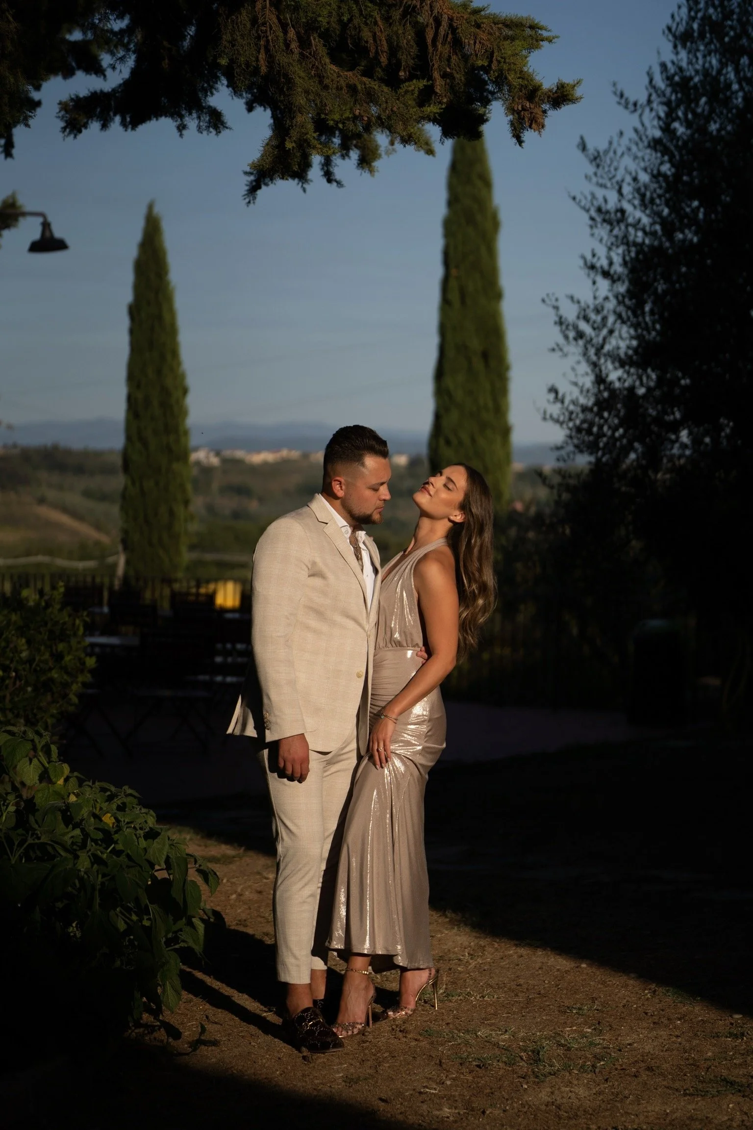 A man and woman standing close together outdoors during dusk, with tall cypress trees and rolling hills in the background. The man wears a light-colored suit, and the woman wears a shiny, metallic gown. They are gazing at each other.