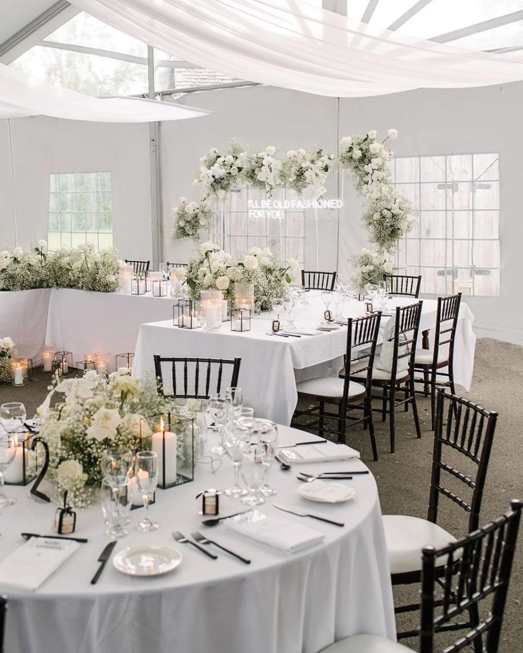Elegant wedding reception setup with white tablecloths, black chairs, floral centerpieces, candles, and a floral arch inside a tent.