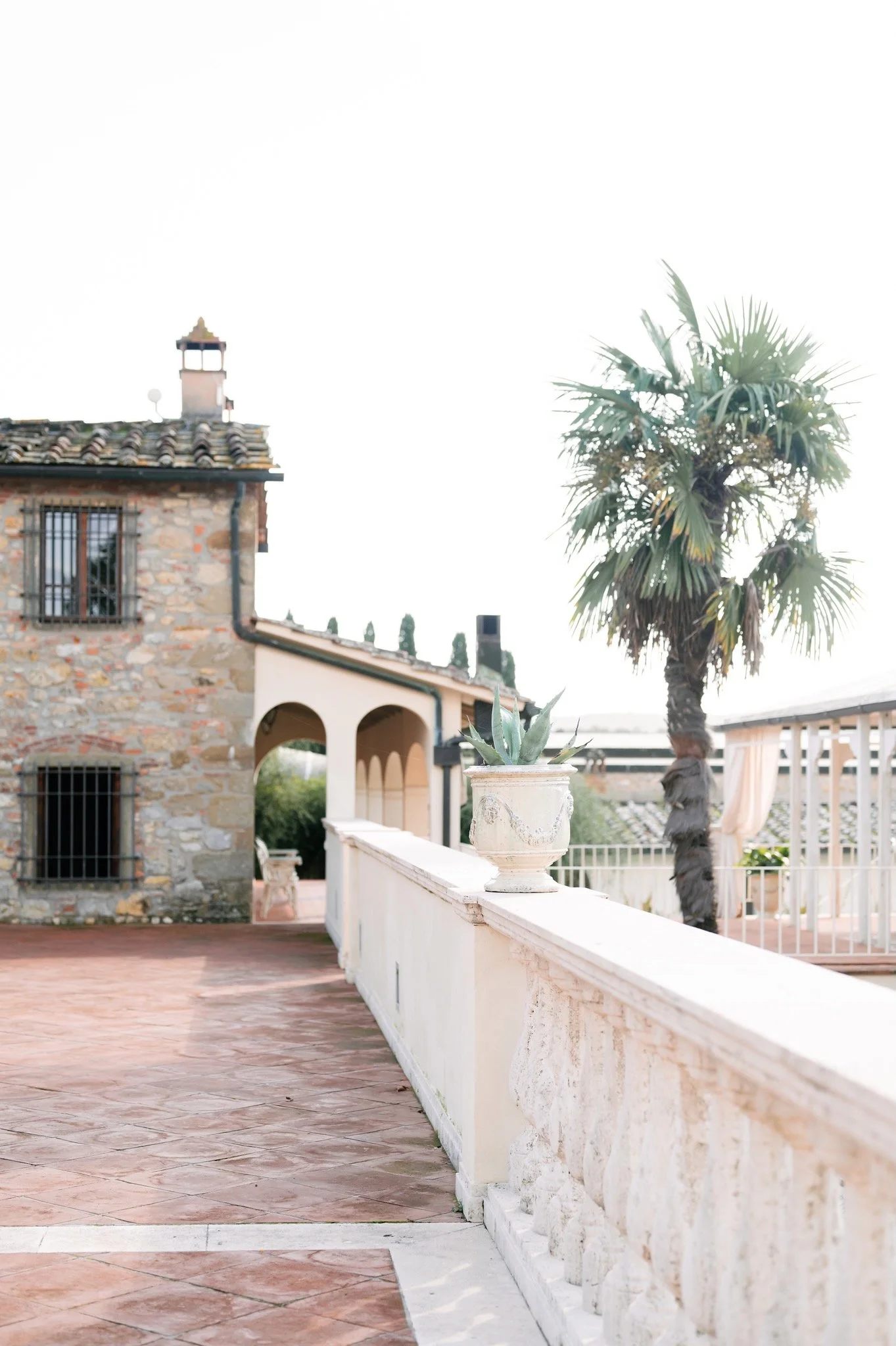 A terrace with terracotta flooring, featuring a stone and stucco building with barred windows, a potted cactus on a ledge, and a tall palm tree with a white railing.