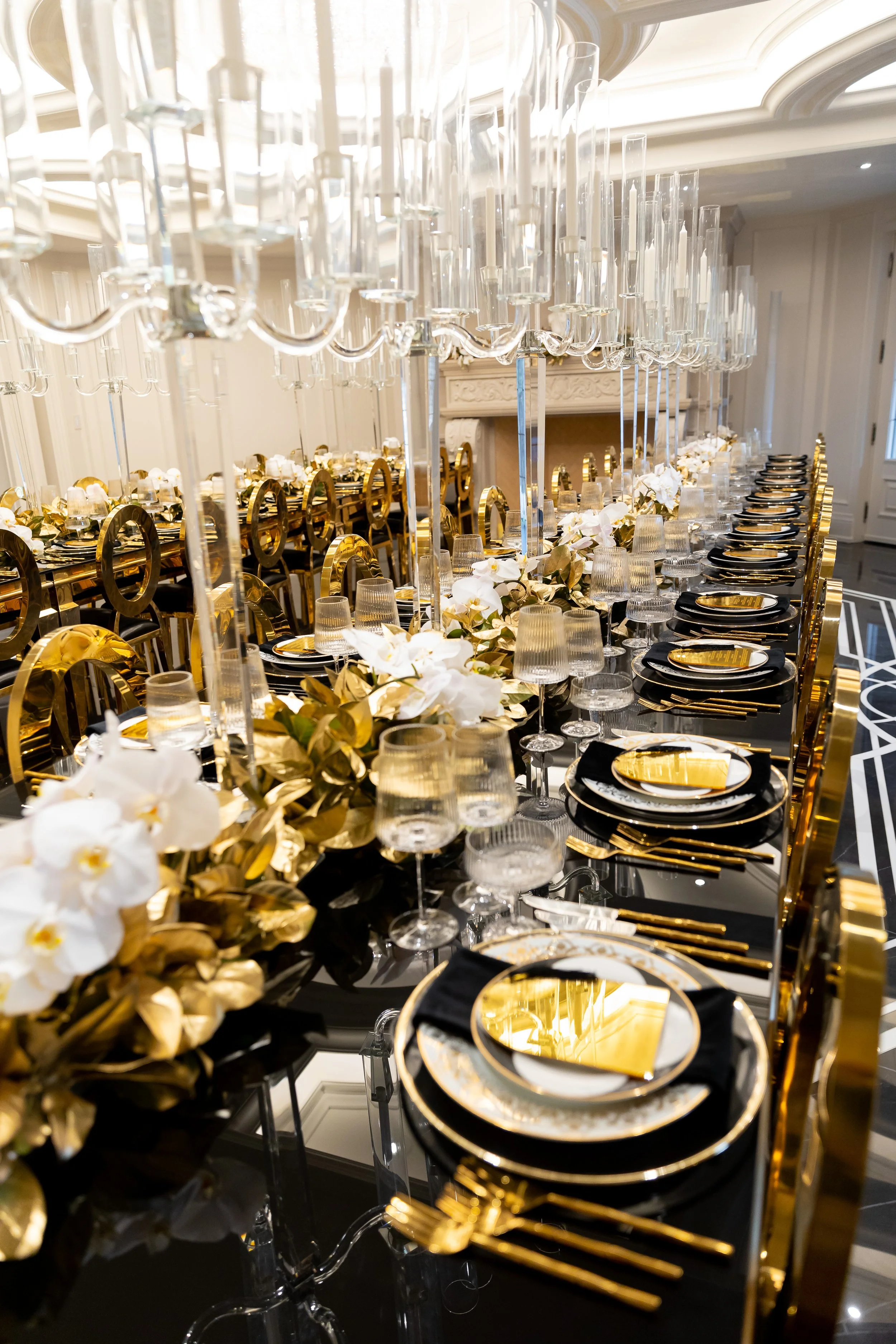Elegant banquet table set with black and gold plates, gold cutlery, and glassware, decorated with white flowers and gold leaves, featuring tall glass candle holders and chandeliers overhead.