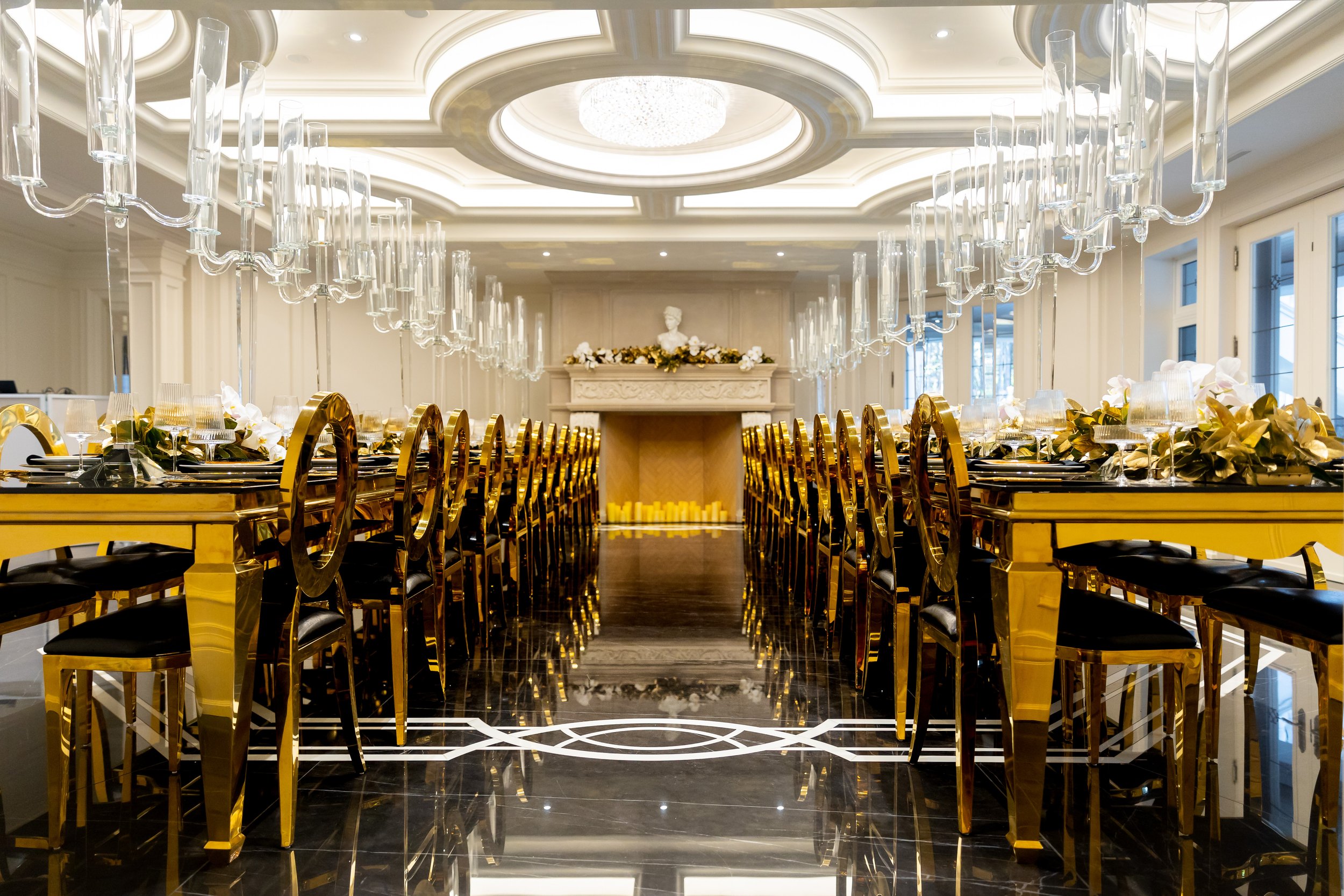 Elegant banquet hall with long gold-trimmed tables, black chairs, glassware, and floral centerpieces, featuring a white fireplace with candles and a classical bust in the background.