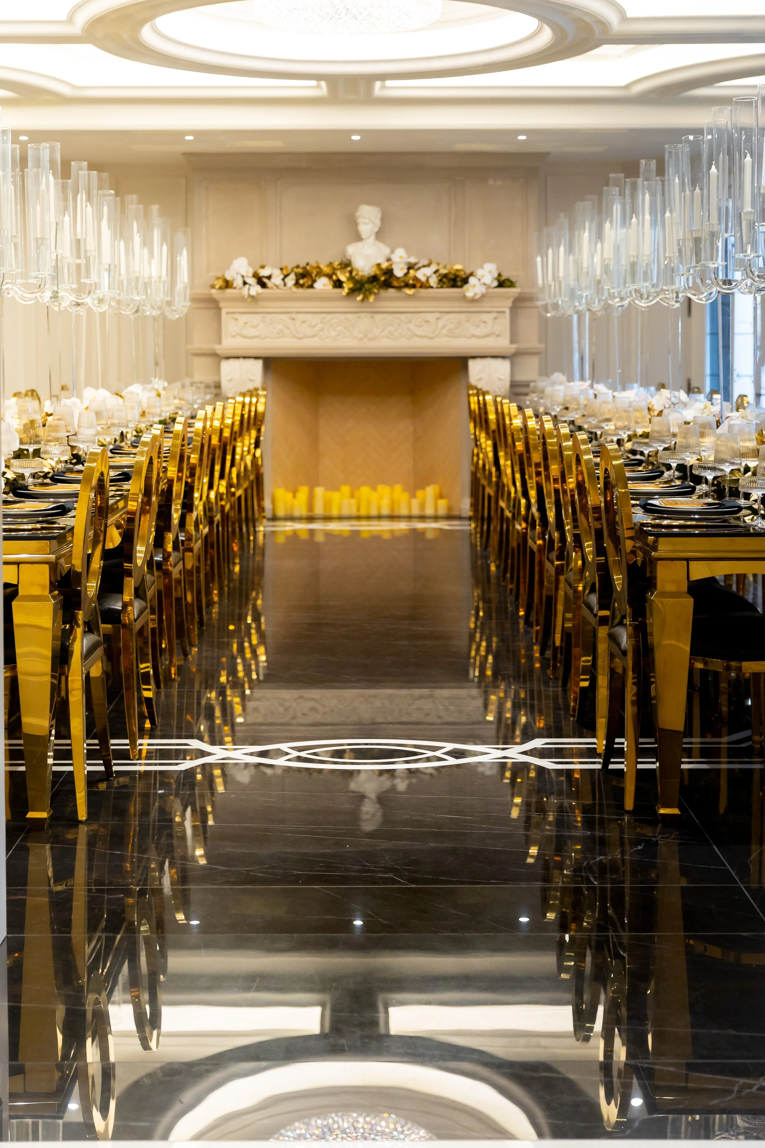A luxurious dining room with a long table set for a formal event, featuring gold chairs, glassware, and a fireplace with candles and a floral mantle.