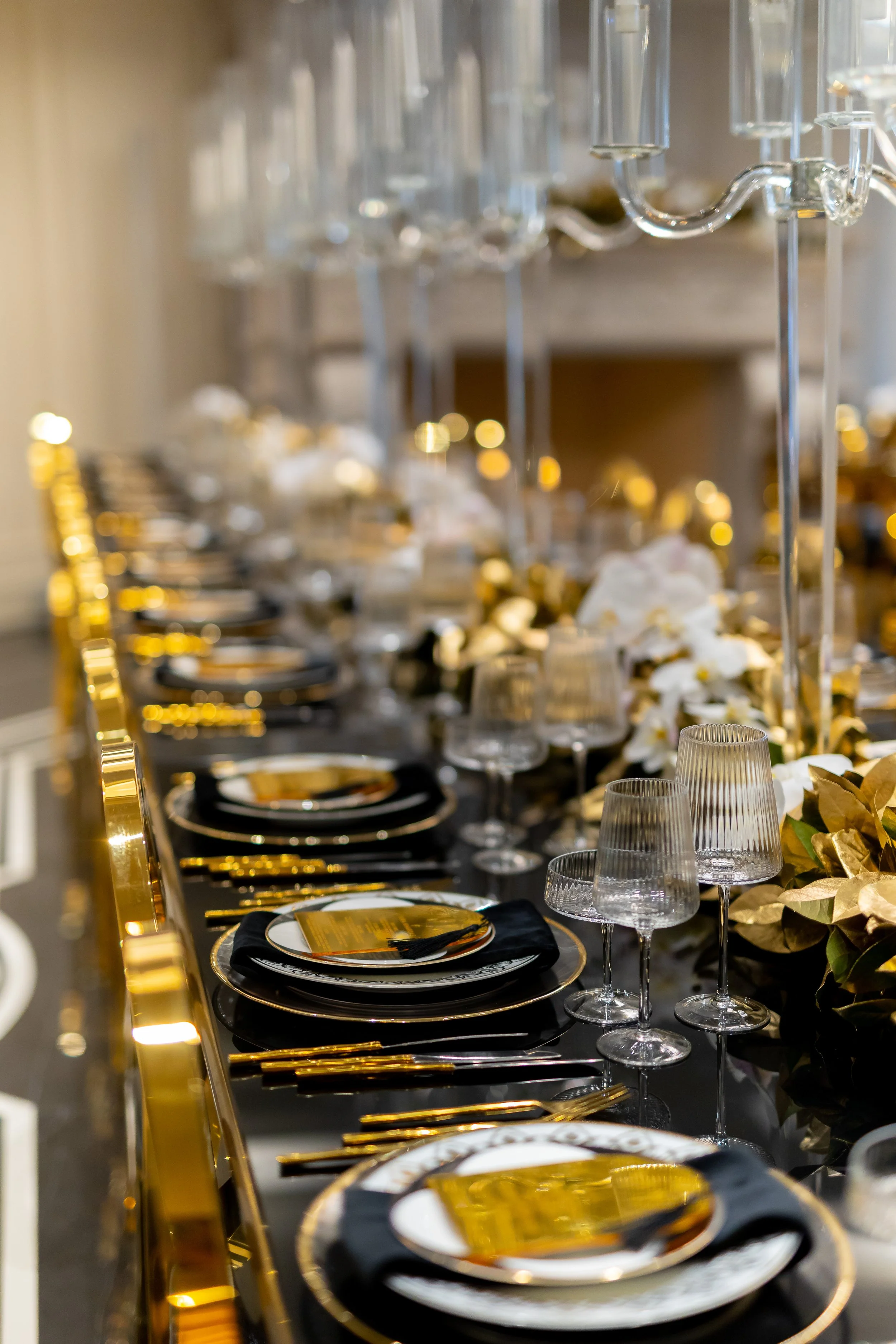 A luxurious banquet table set with black and gold dinnerware, gold utensils, clear glassware, and floral arrangements with white flowers and gold leaves.