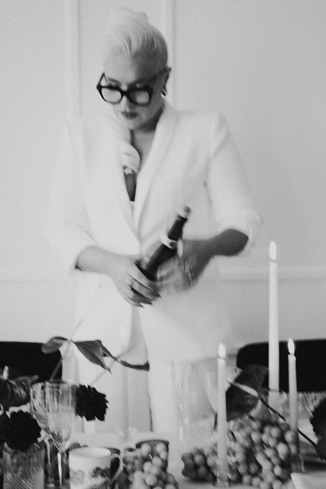 A woman with blonde hair, glasses, and a white blazer holding a bottle of champagne in a room decorated with candles, flowers, and baskets of fruit on a table.
