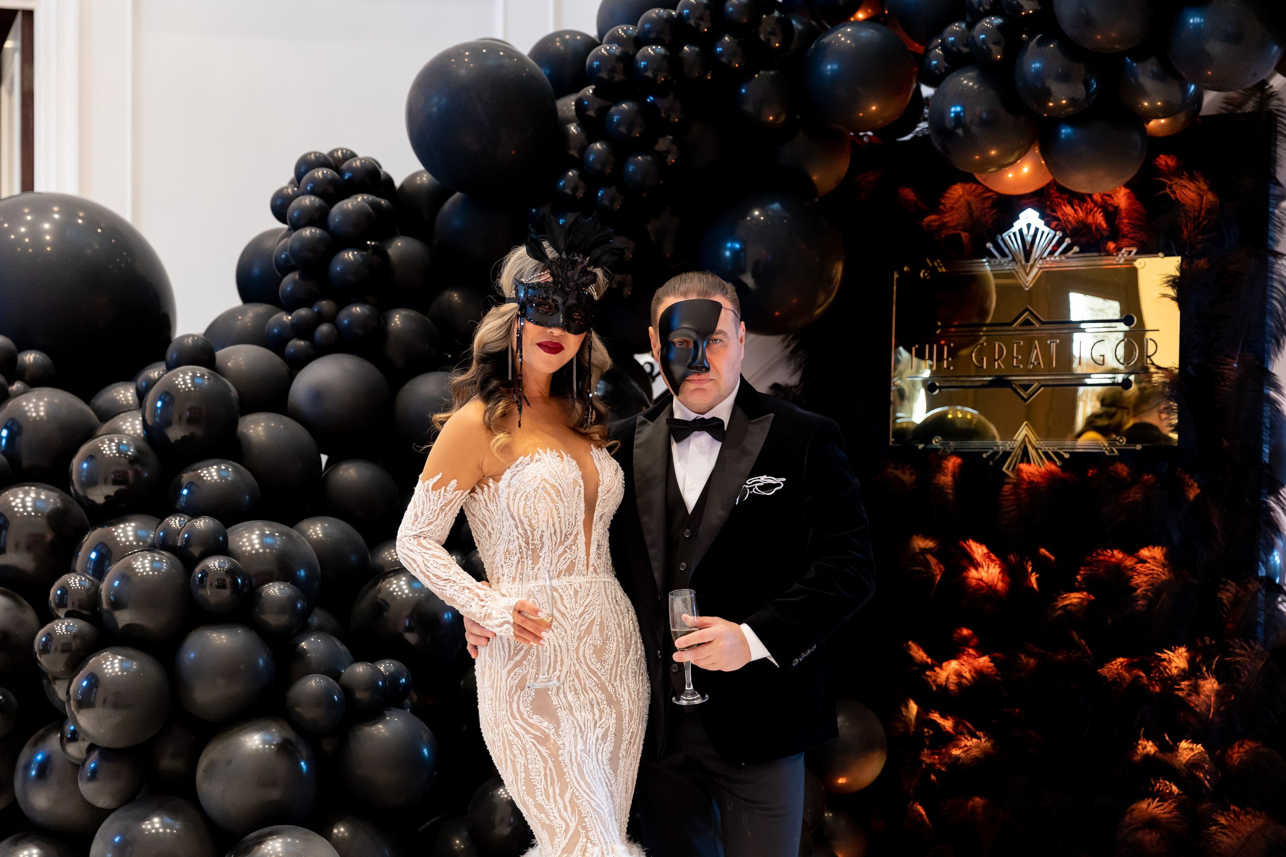 A man and woman dressed in formal attire, wearing decorative masks, standing close together at a black and gold themed event with black balloons and a sign that reads 'ONE: GREAT GOD' in the background.