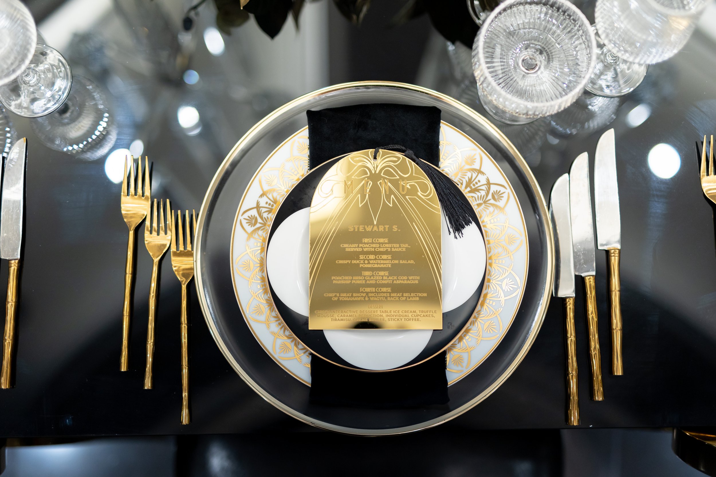 A formal dining table setting with gold cutlery, a black and white patterned plate, a black napkin, and a decorative menu card with gold details.